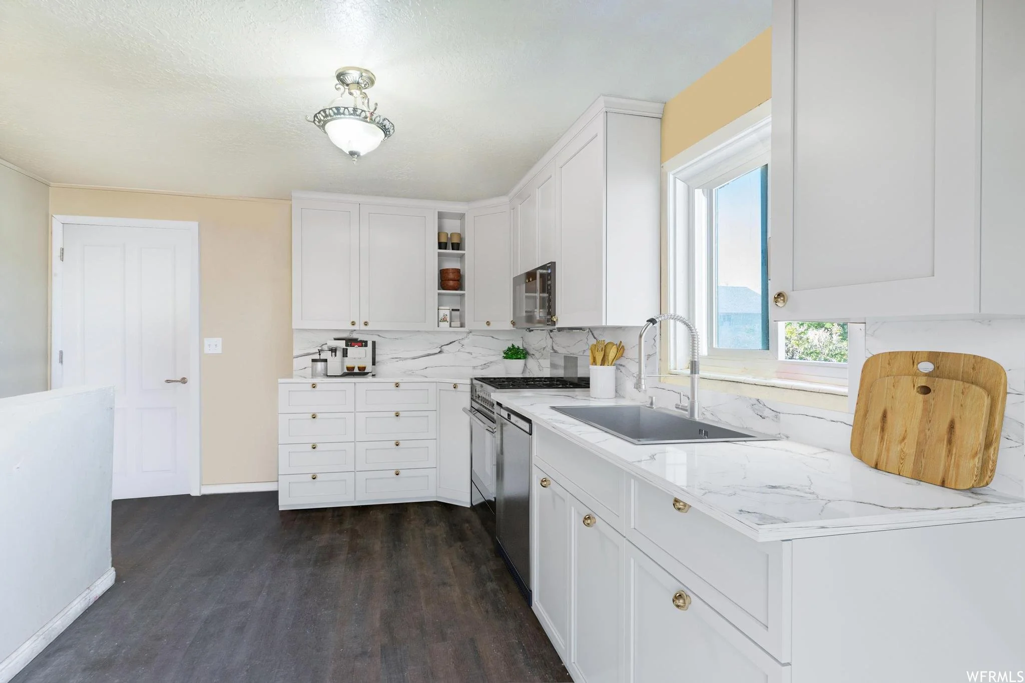 Modern kitchen with white cabinets, marble countertops, stainless steel appliances, and dark wood flooring. Features a sink, cutting board, and built-in microwave.