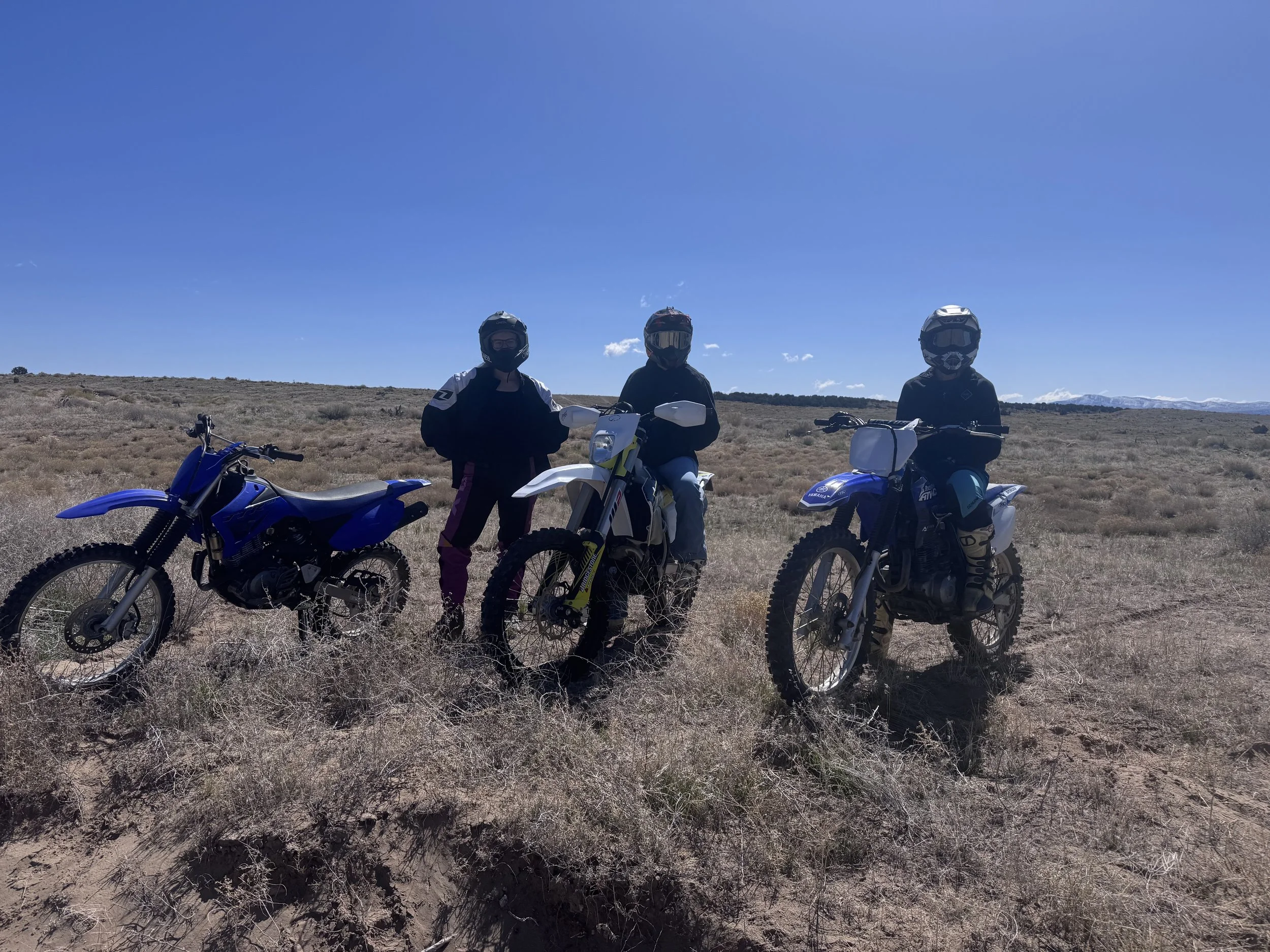 Three people in motorcycle gear standing next to dirt bikes in a dry, open landscape under a clear blue sky.