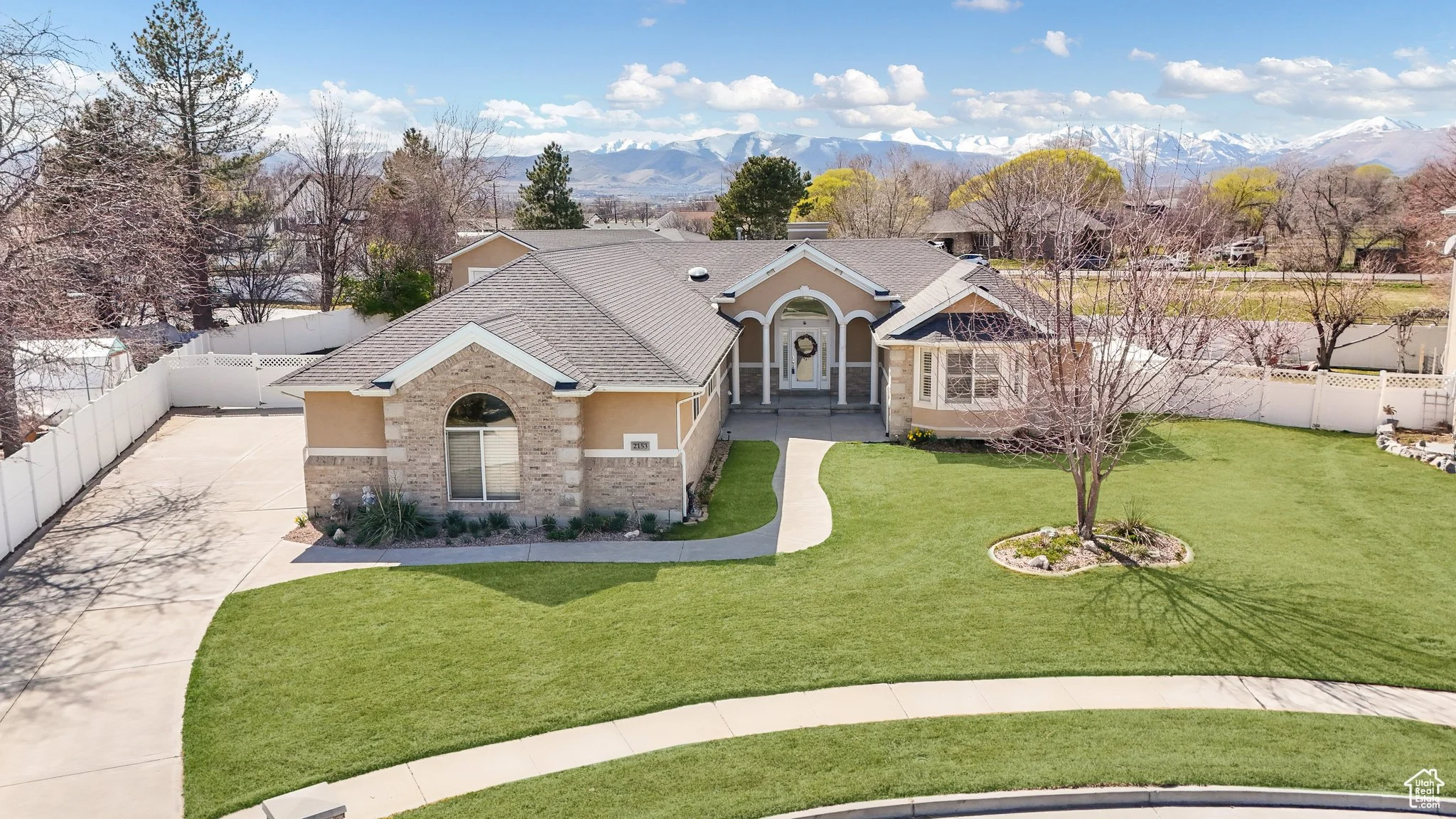 A suburban house with a brick facade, arched windows, and a well-maintained front lawn. The driveway is paved, and there is a path leading to the entrance. Trees with bare branches surround the property, with a backdrop of distant mountains under a partly cloudy sky.