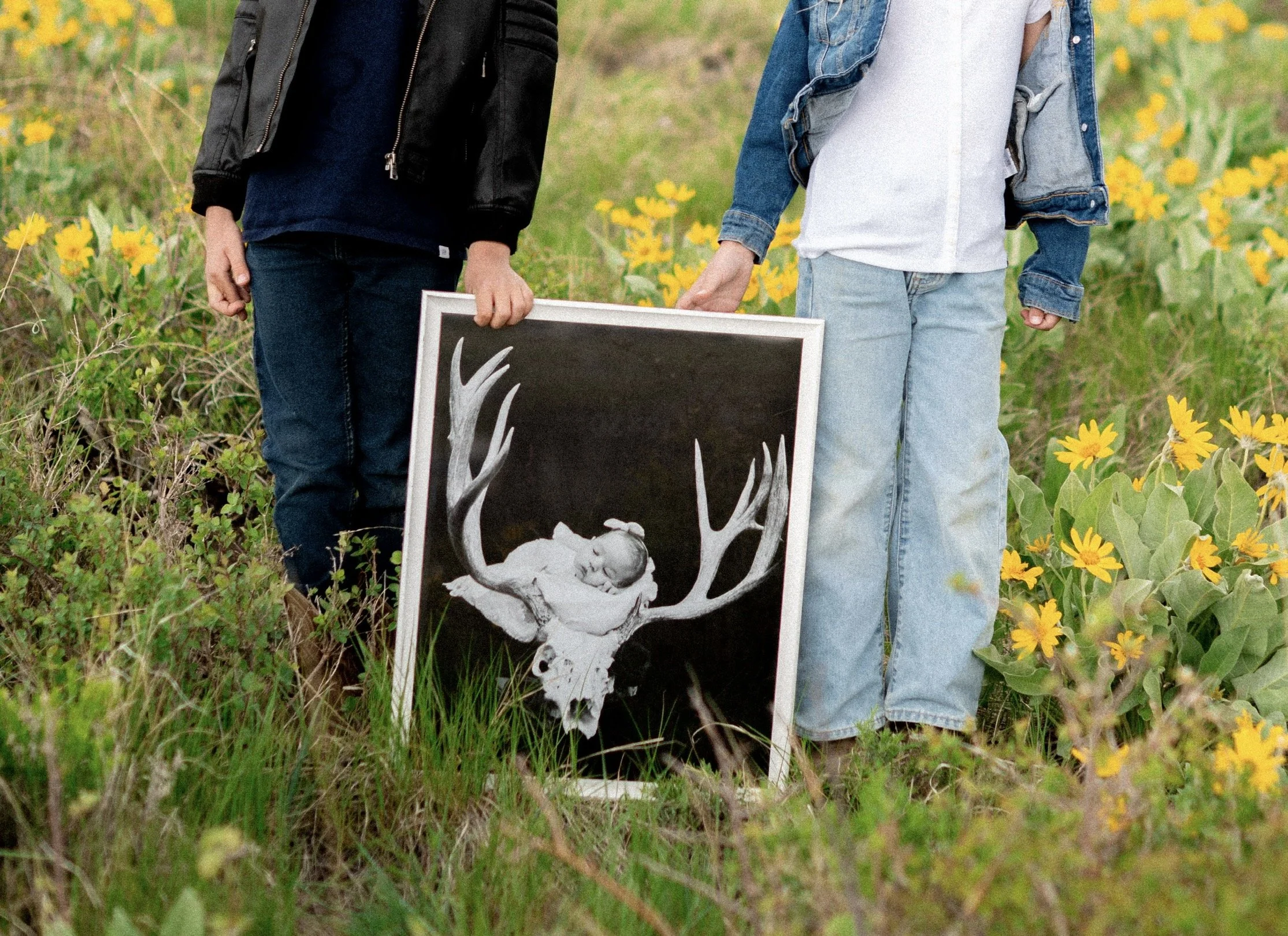 Two children holding a framed black and white photo of a baby resting on antlers, standing in a field with yellow flowers.