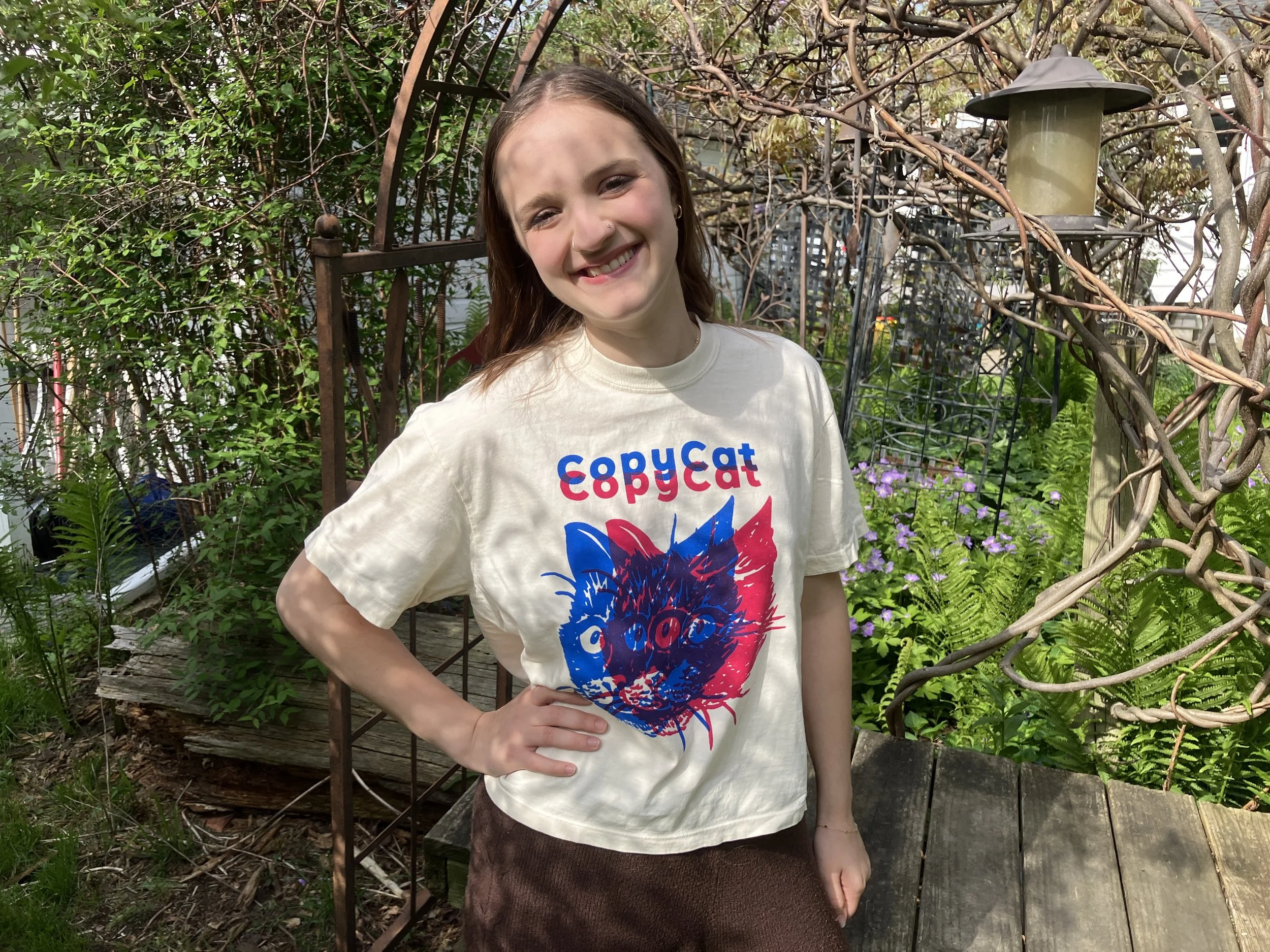 A person wearing a cropped, off-white CopyCat t-shirt which features overlapping illustrated cat heads in pink and blue.