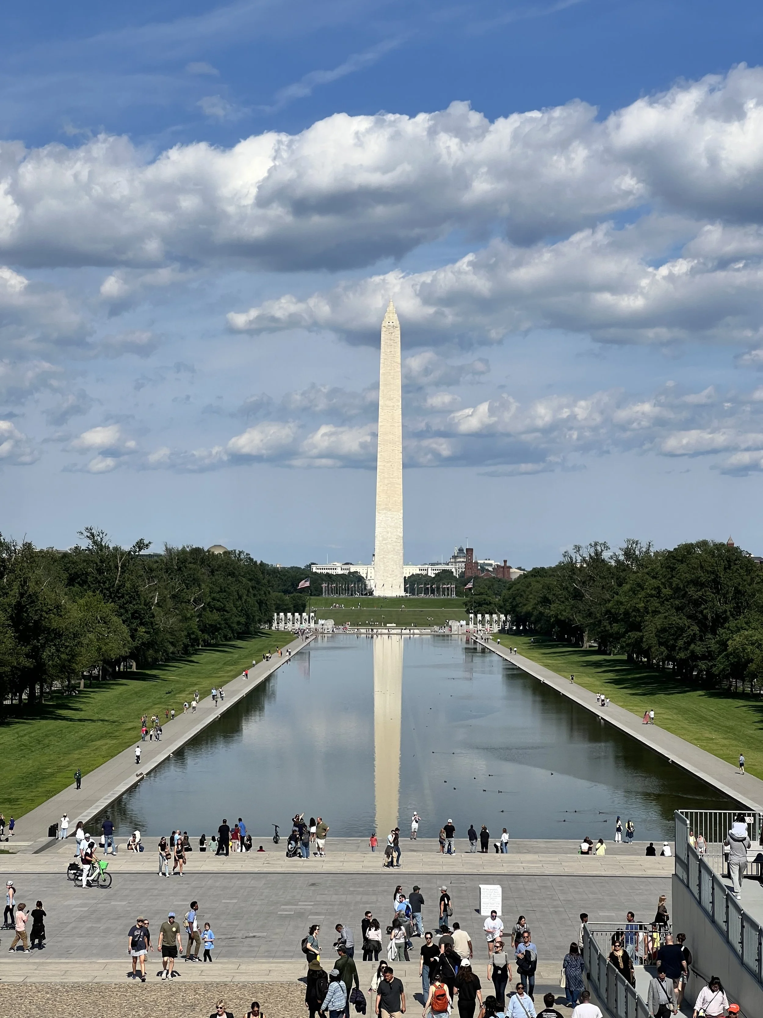 Le reflet de l'obélisque de Washington dans le miroir d'eau du National Mall à Washington, D.C., avec des gens se promenant alentour et des nuages dans le ciel.