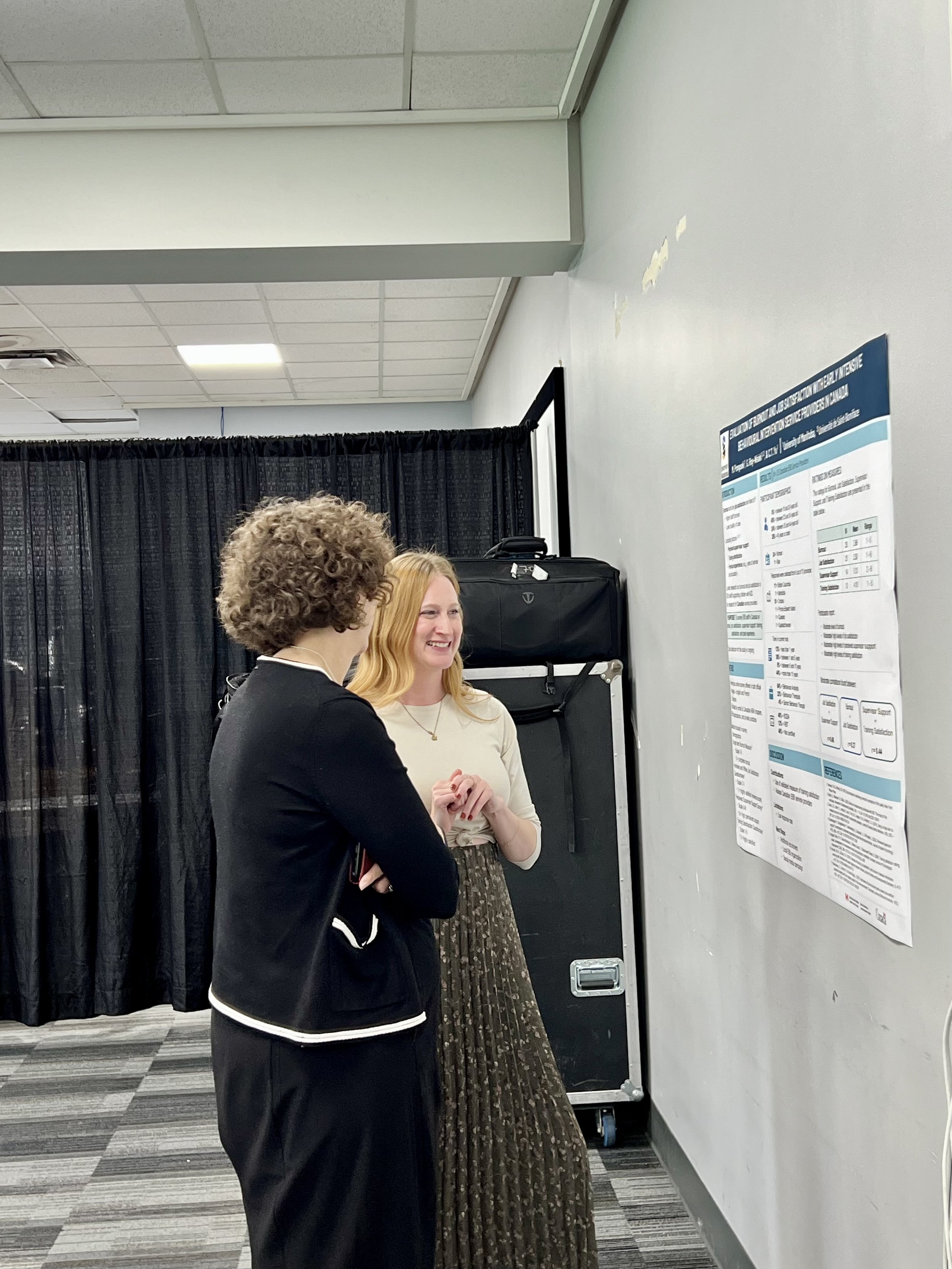 Deux femmes discutent devant un poster scientifique affiché sur un mur blanc dans une salle de conférence.