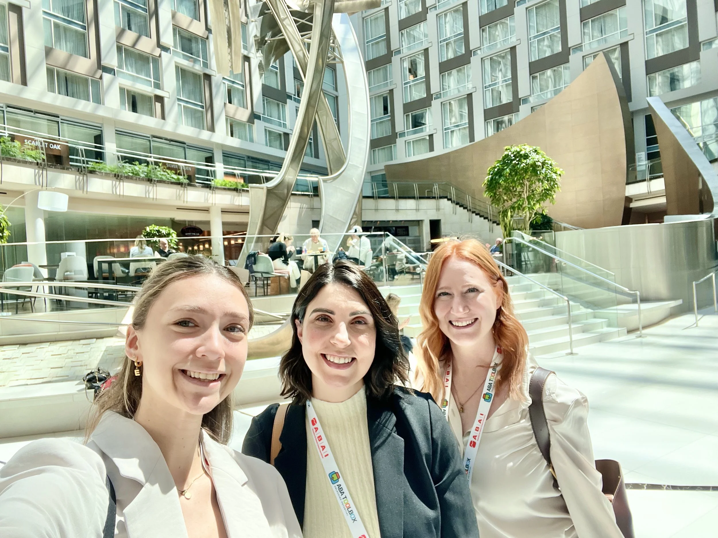 Trois femmes souriantes prennent une photo dans un bâtiment moderne, avec des escalators, des plantes vertes et des personnes en arrière-plan.