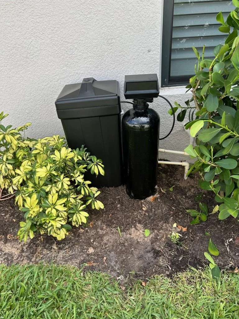 Outdoors near a house wall, a black brine tank, a black water softener, and green bushes on soil and grass.