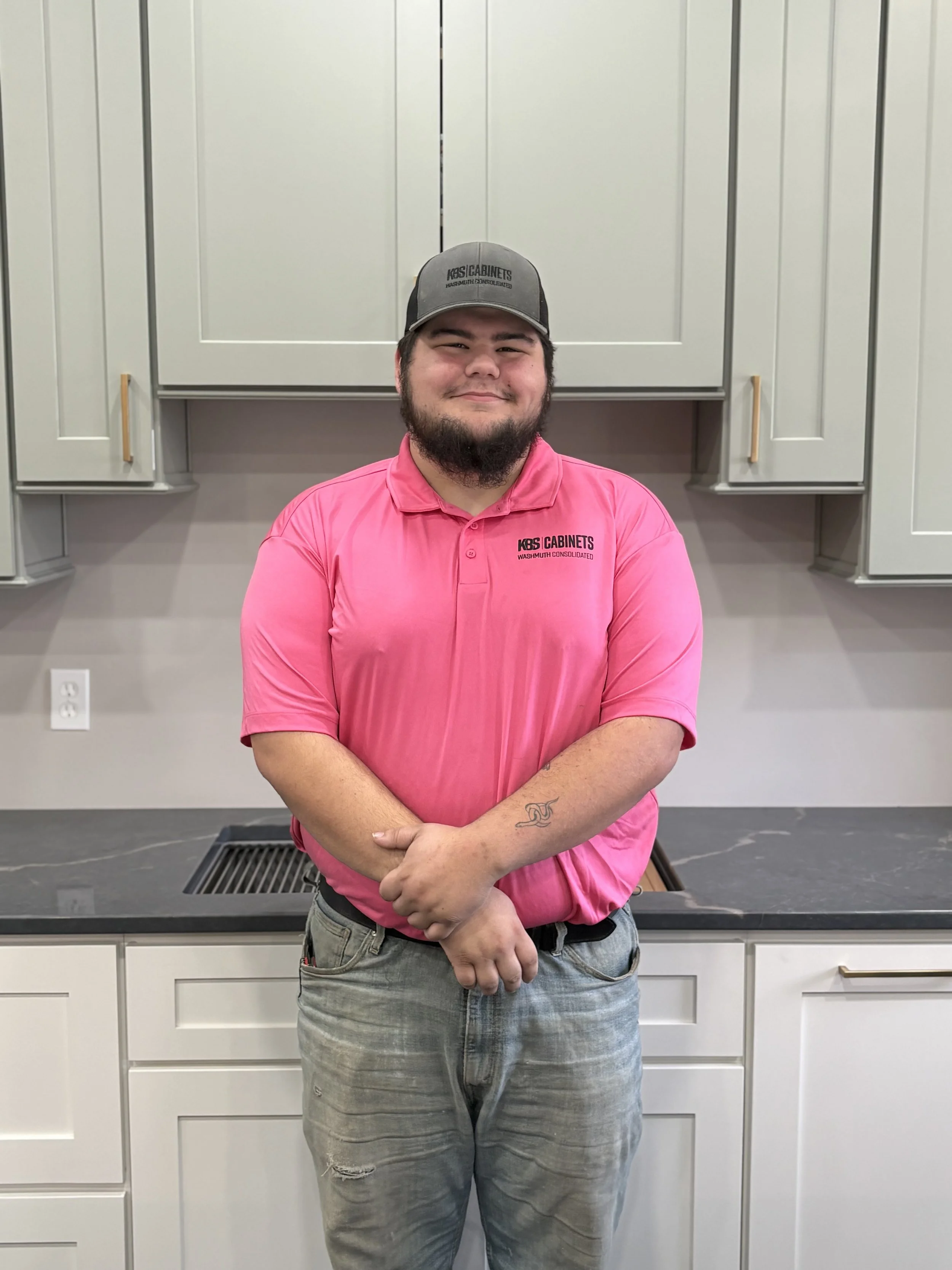 A young man with a beard, wearing a black cap, gray t-shirt, and light jeans, standing indoors in front of kitchen cabinets and appliances, smiling at the camera.