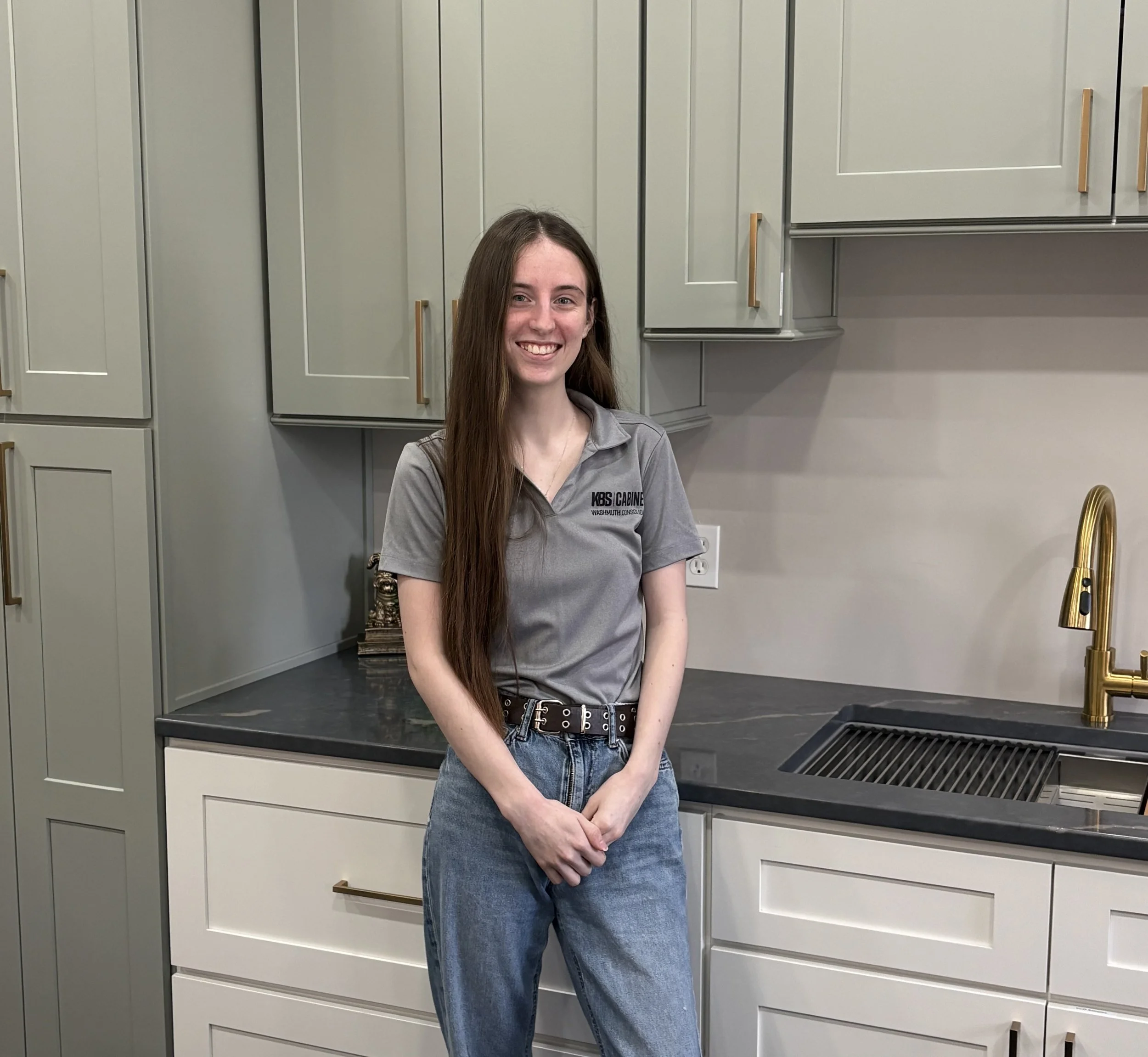 A young woman with long brown hair and blue eyes, smiling, wearing a dark hoodie, standing in front of a textured stone wall.