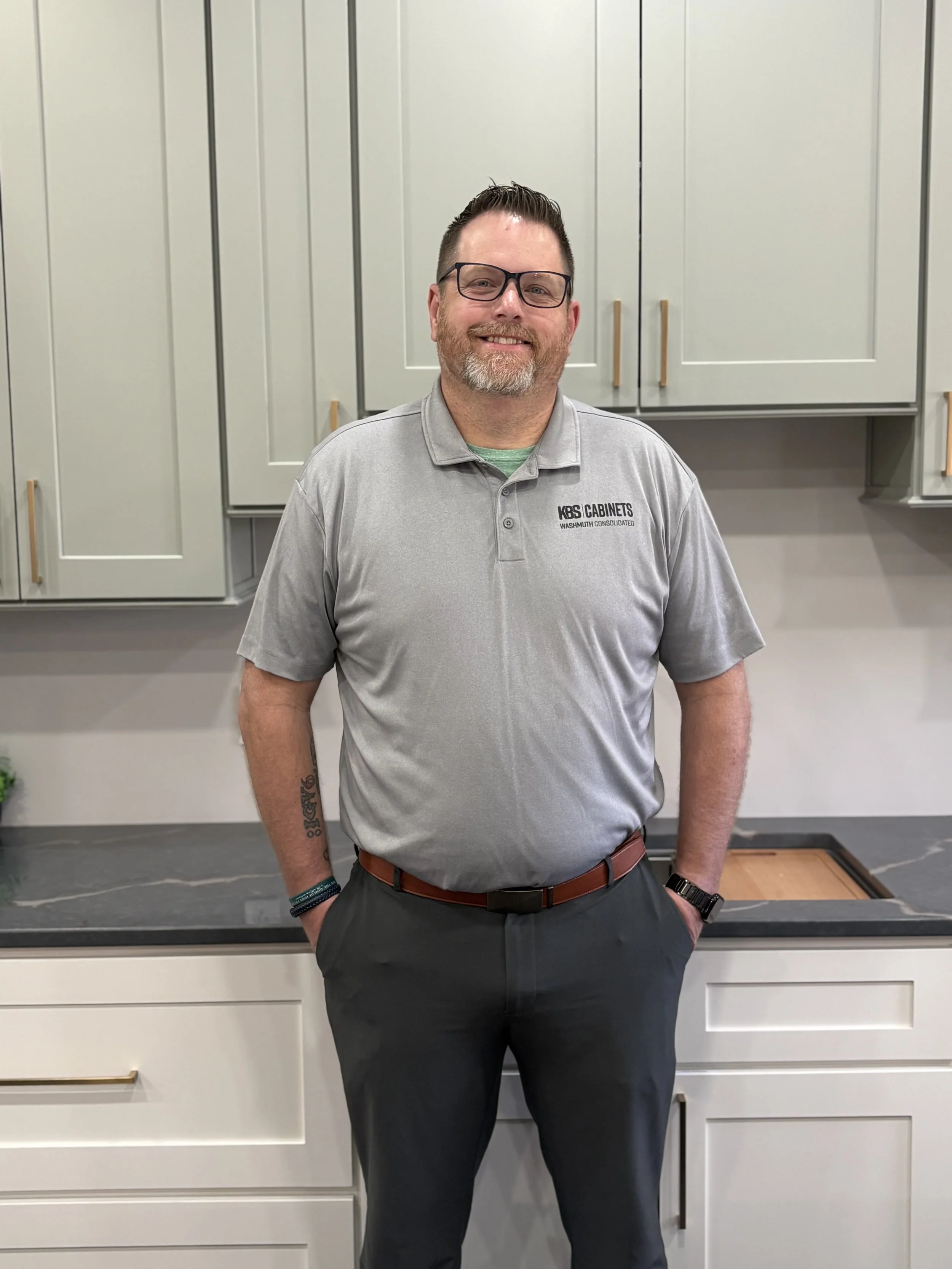A man with glasses, a beard, and tattoos on his arms standing with arms crossed in a kitchen with green cabinets, a coffee maker, potted plants, and dog figurines on the counter.