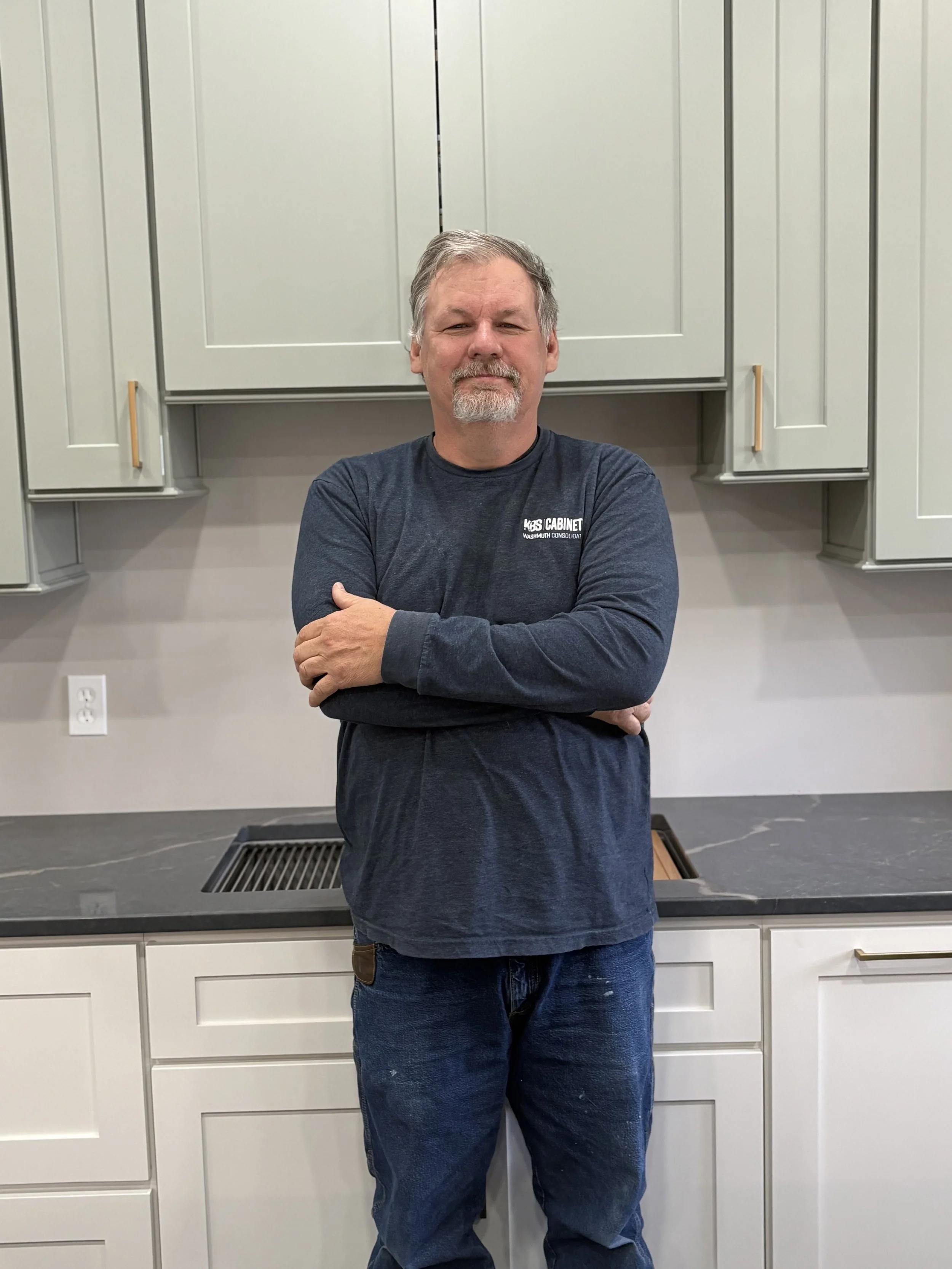 A man with glasses, gray hair, and a beard standing in a kitchen with arms crossed, wearing a light blue t-shirt and dark pants.