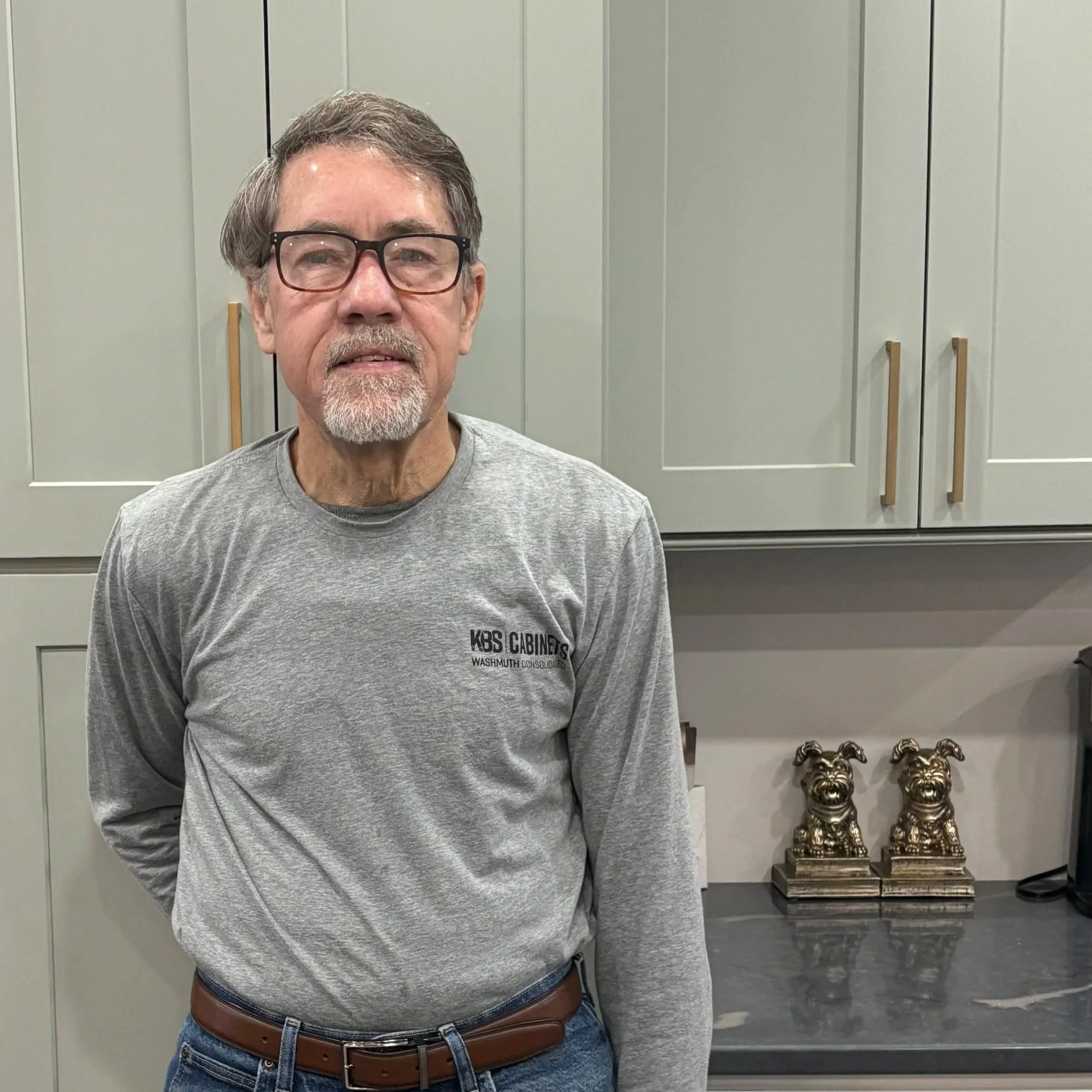 An older man with glasses and a beard and mustache, wearing a gray long-sleeve shirt and jeans, standing in a kitchen with gray cabinets and two dog statues on the counter.