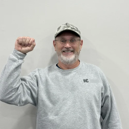 A smiling man with glasses, a gray beard, and a gray sweatshirt with 'WC' embroidered on it, raising his right fist in a gesture of victory or celebration against a plain white wall.