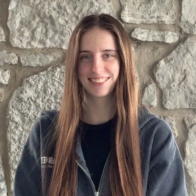 A young woman with long brown hair and blue eyes, smiling, wearing a dark hoodie, standing in front of a textured stone wall.