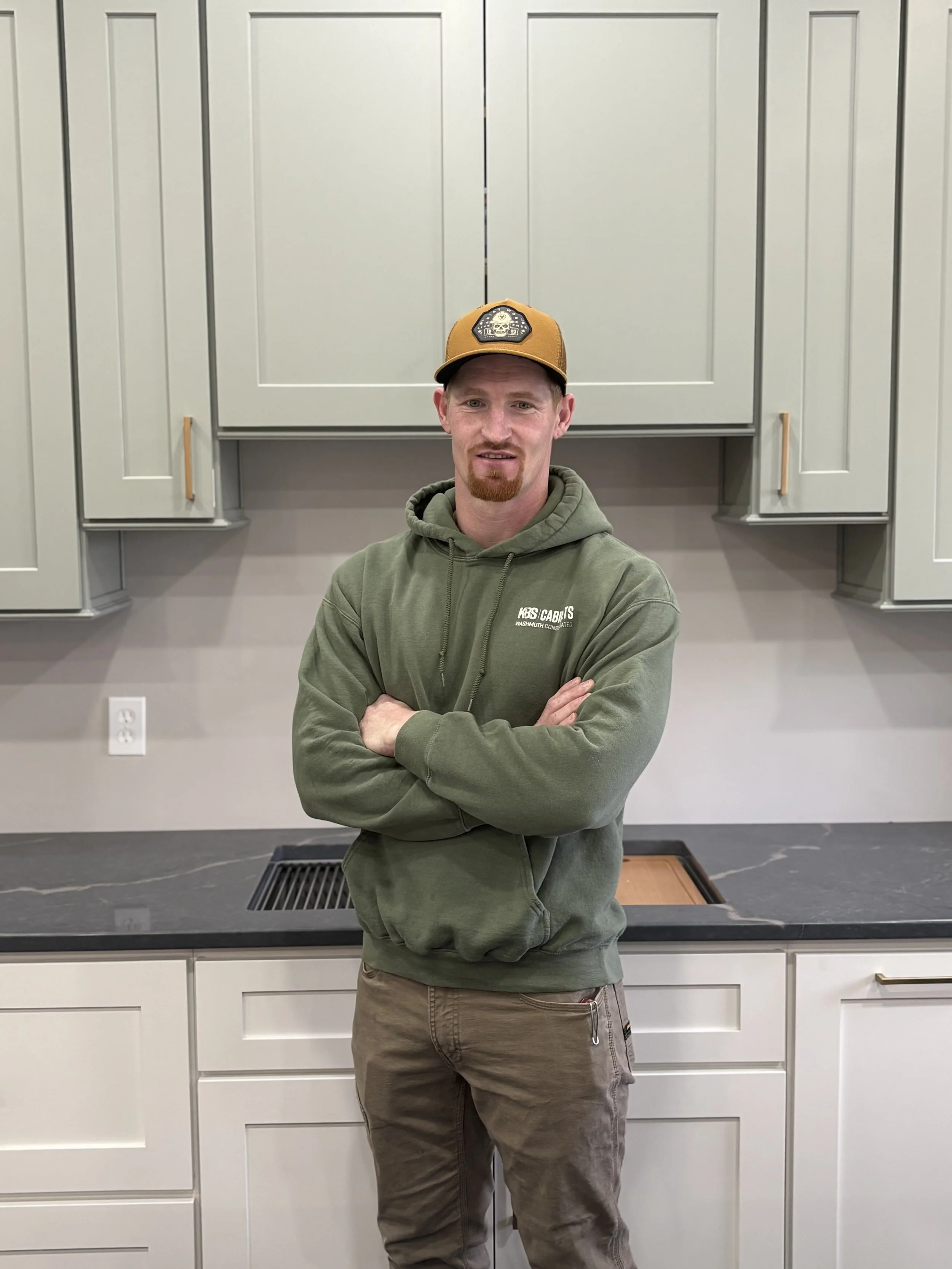 A man wearing a light gray baseball cap, an olive green hoodie with 'KBS Cabinets' logo, and khaki pants standing in front of wooden kitchen cabinets with a light-colored countertop and a small potted plant.