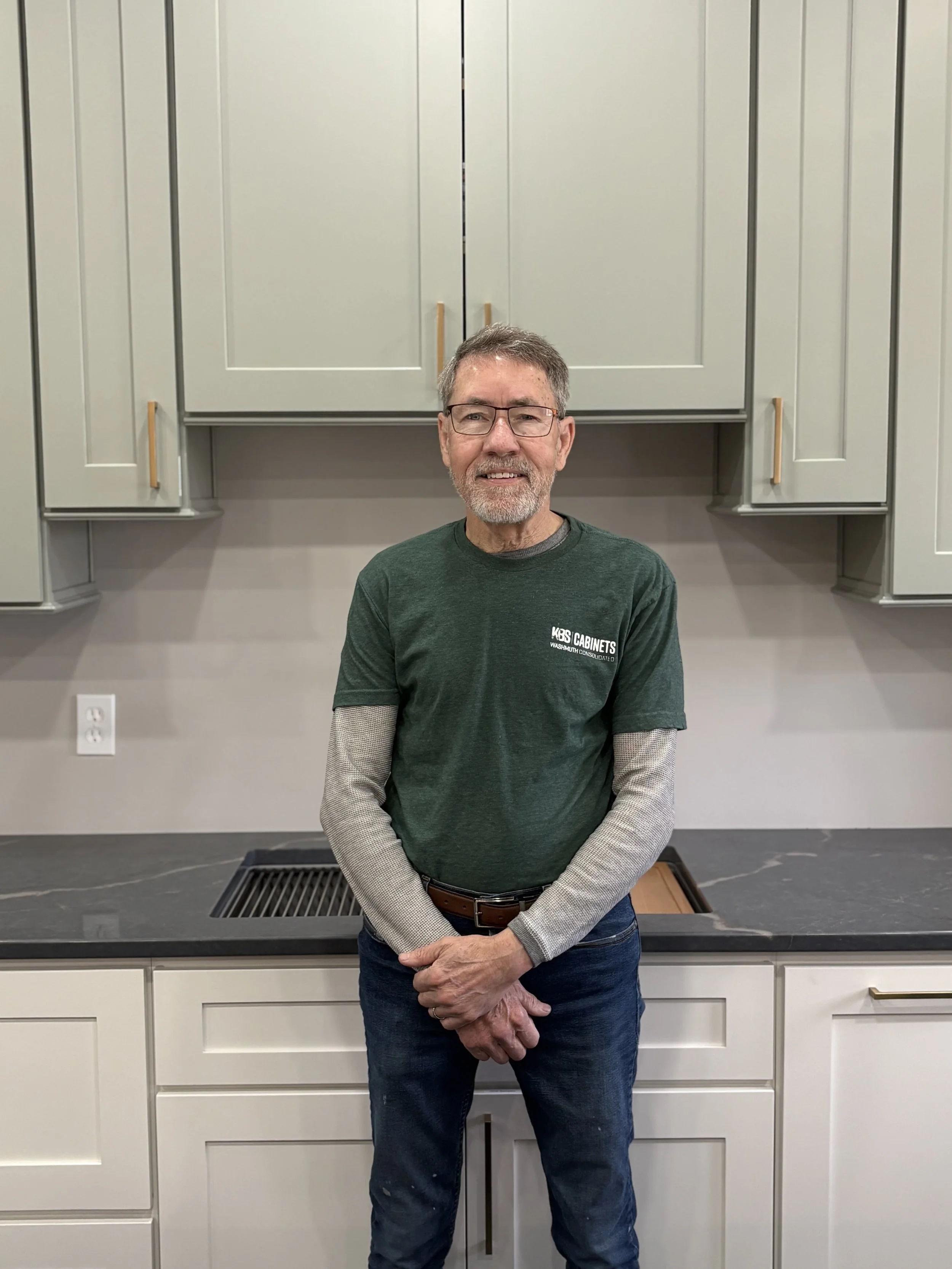 An older man with glasses and a beard and mustache, wearing a gray long-sleeve shirt and jeans, standing in a kitchen with gray cabinets and two dog statues on the counter.