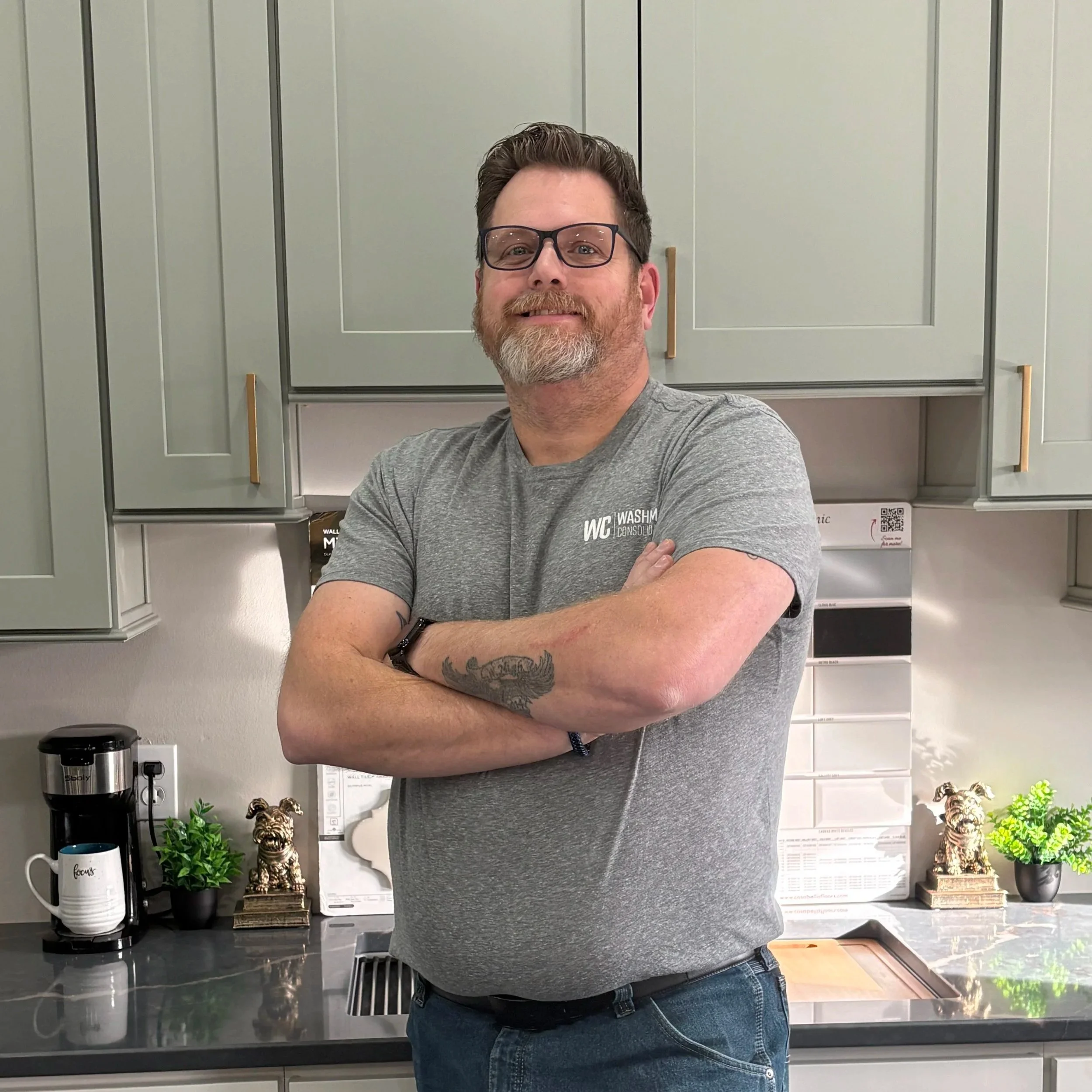 A man with glasses, a beard, and tattoos on his arms standing with arms crossed in a kitchen with green cabinets, a coffee maker, potted plants, and dog figurines on the counter.