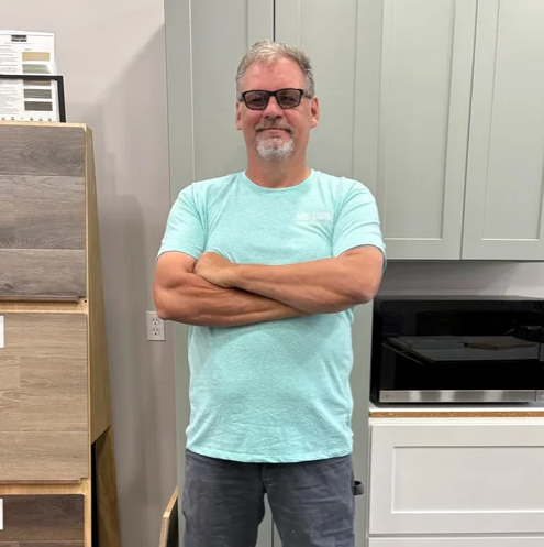 A man with glasses, gray hair, and a beard standing in a kitchen with arms crossed, wearing a light blue t-shirt and dark pants.