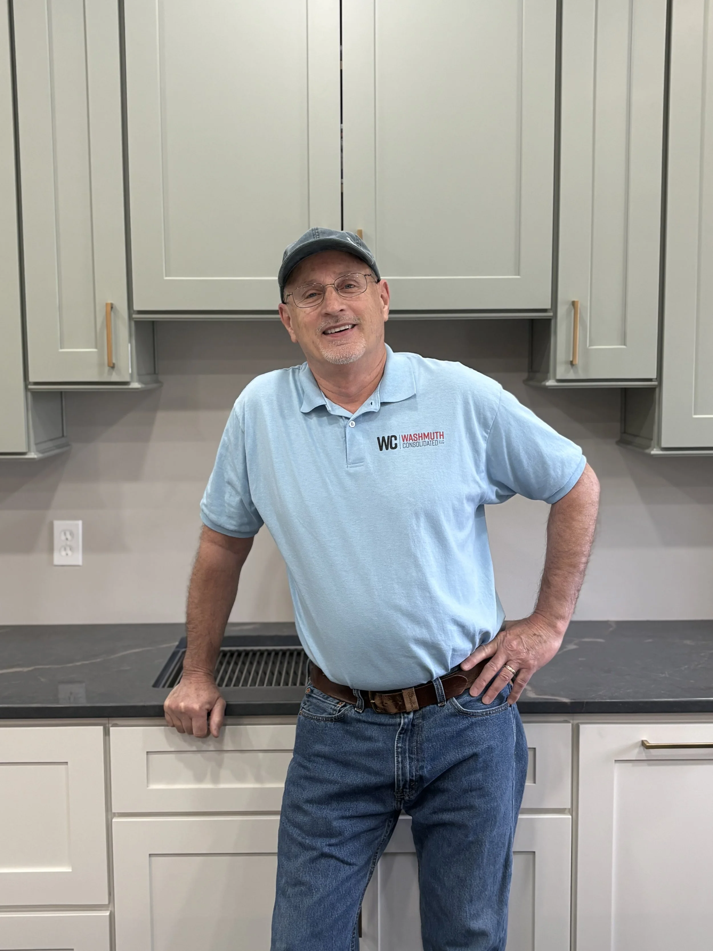 A smiling man with glasses, a gray beard, and a gray sweatshirt with 'WC' embroidered on it, raising his right fist in a gesture of victory or celebration against a plain white wall.