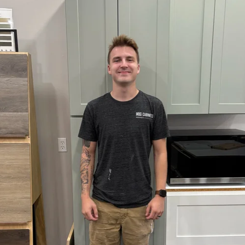 A young man standing in a kitchen, smiling at the camera. He is wearing a dark T-shirt, khaki shorts, and a smartwatch.