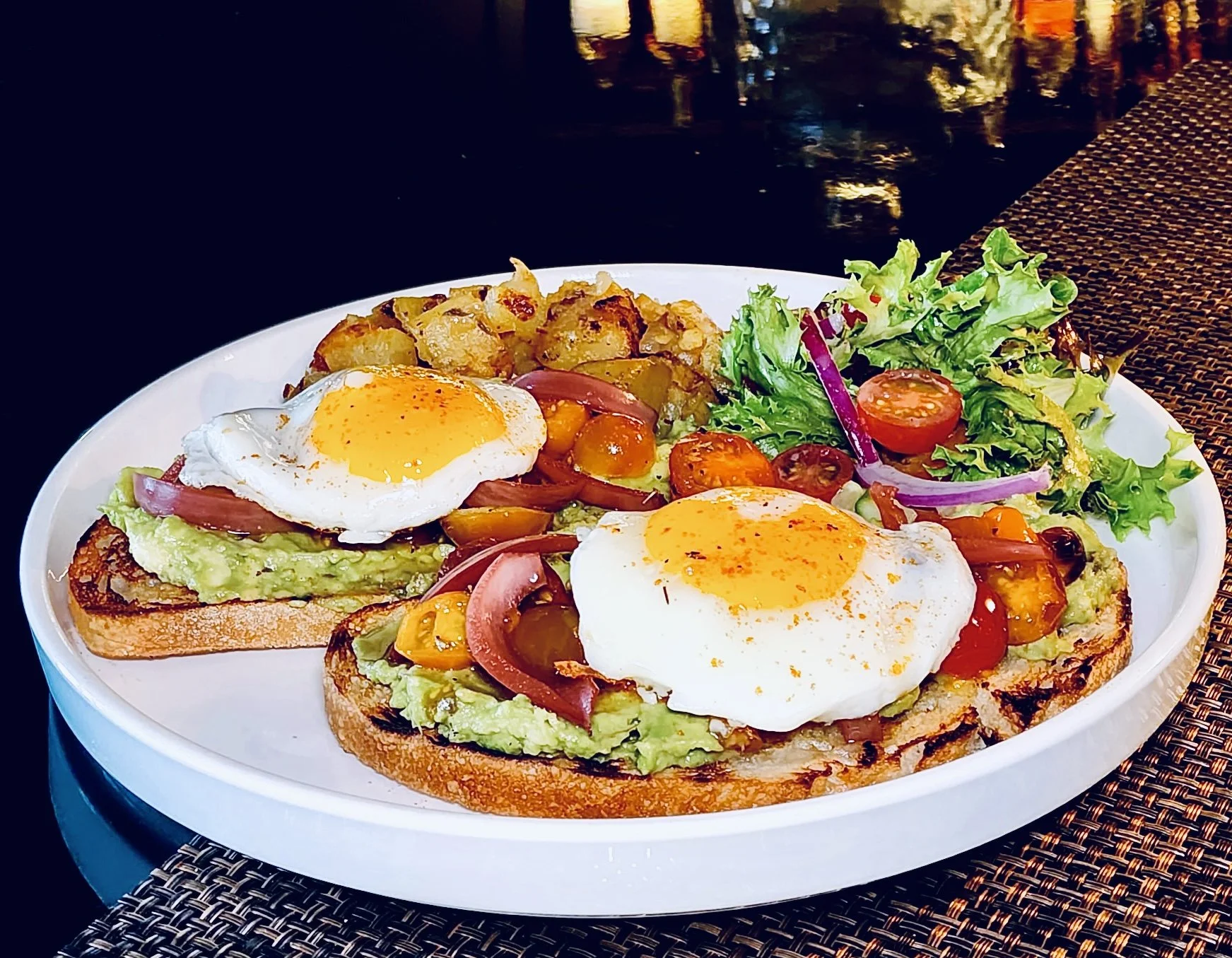 Open-faced avocado, tomato, and onion toast with fried eggs, atchara, and a side salad on a white plate.