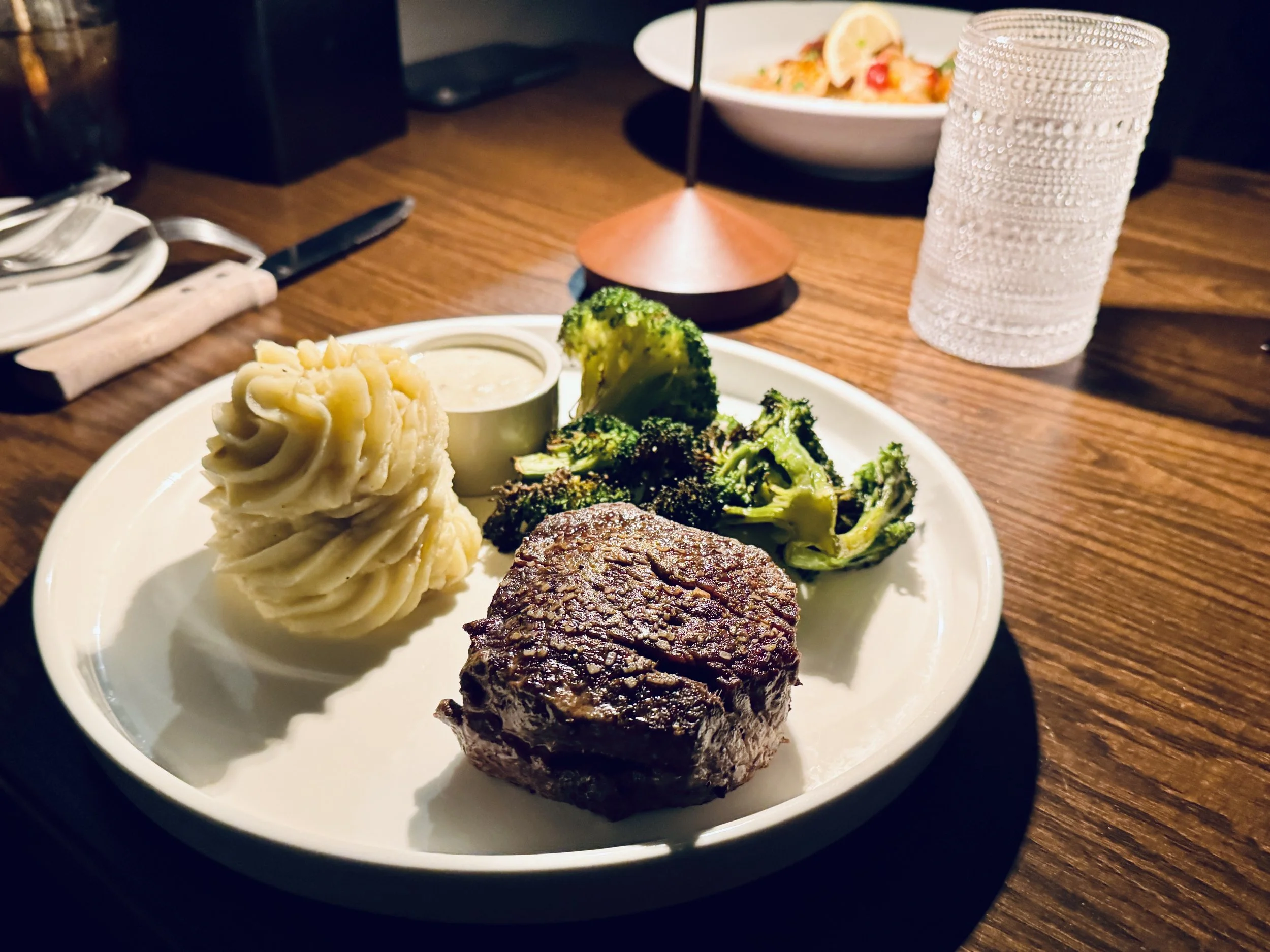 Plate with mashed potatoes, broccoli, grilled steak, and a small cup of sauce on a wooden table in a restaurant setting.