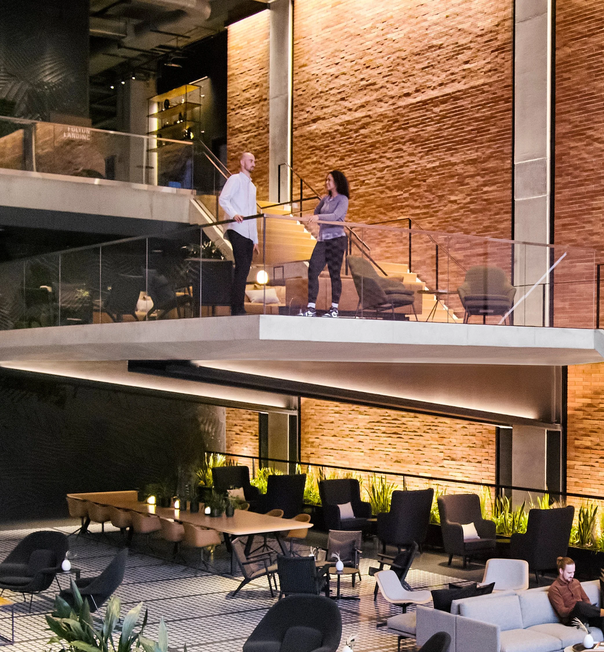 Two people standing and talking on a glass-railed balcony in a modern, layered interior space with warm lighting, exposed brick walls, and contemporary furniture, including armchairs and tables.