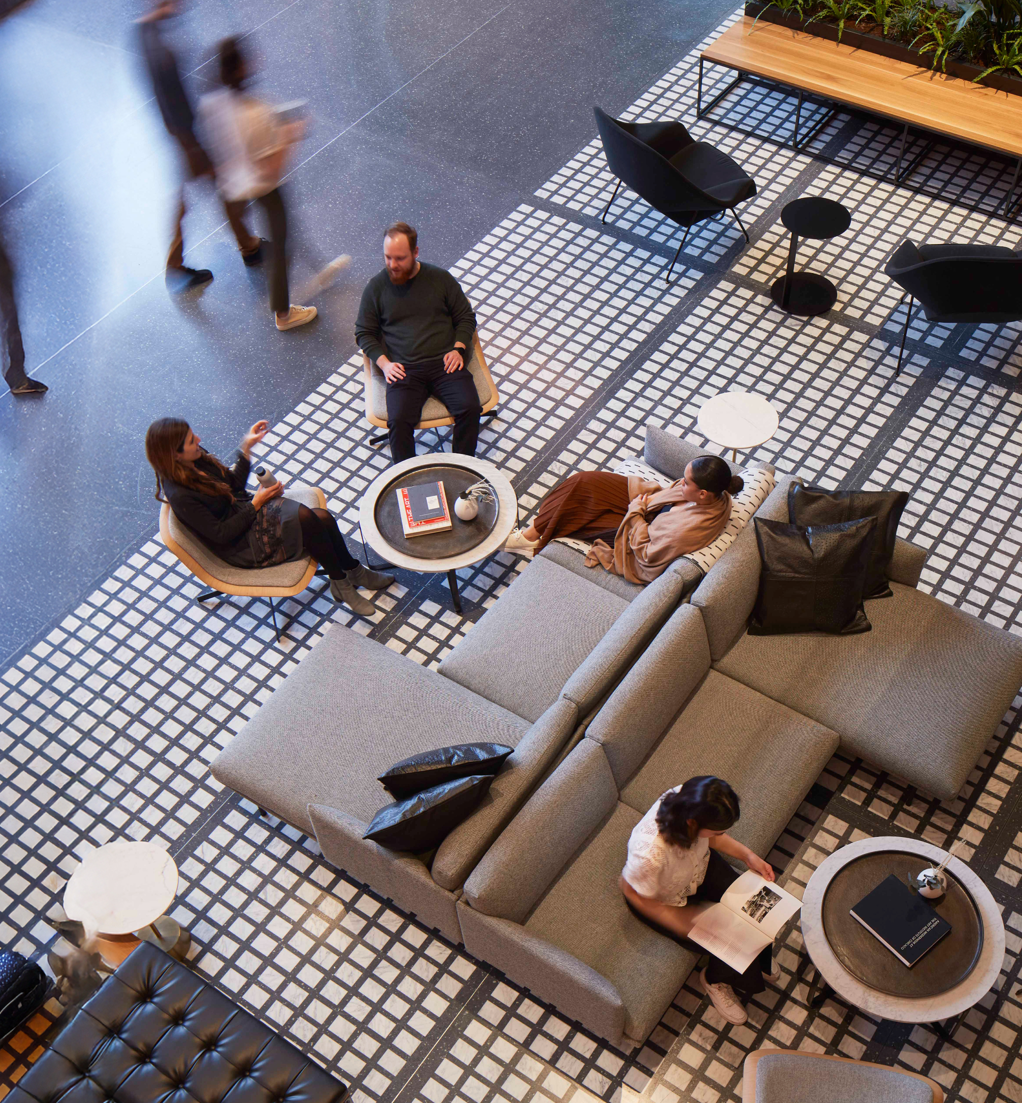 People sitting and talking in a modern, stylish lounge area with black and white tiled flooring, gray sofa, armchairs, and small tables with books and decorative items.