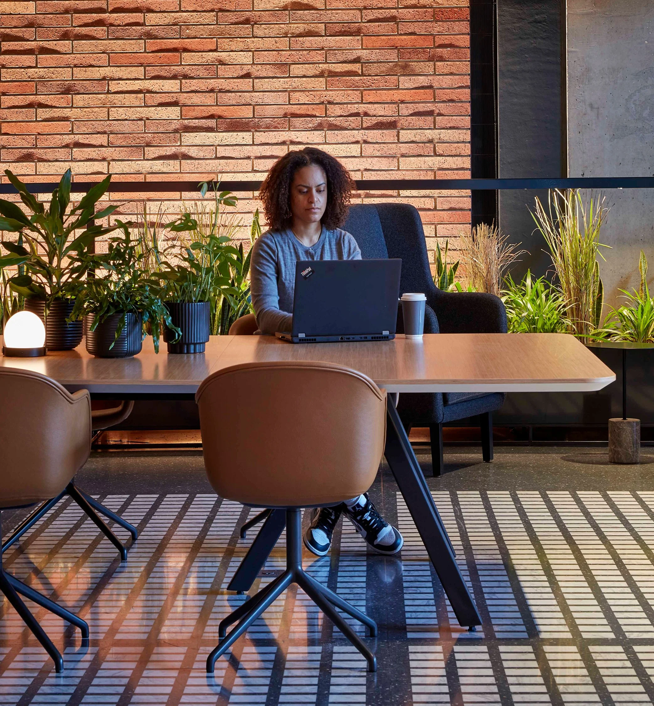 A woman working on a laptop at a wooden table in a modern cafe or office with a brick wall and green plants in the background.