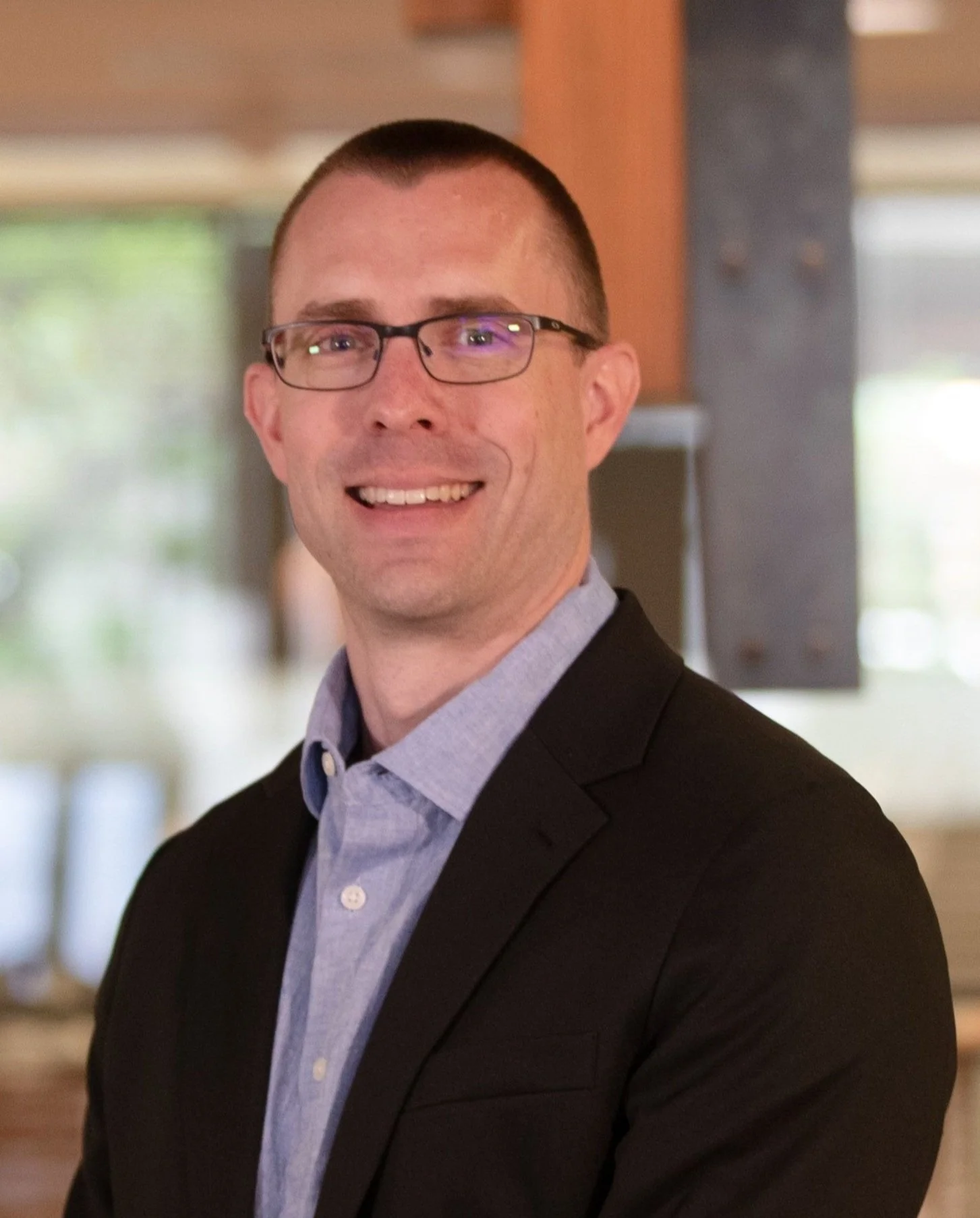 A man in glasses wearing a dark suit jacket and a light blue shirt posing indoors.