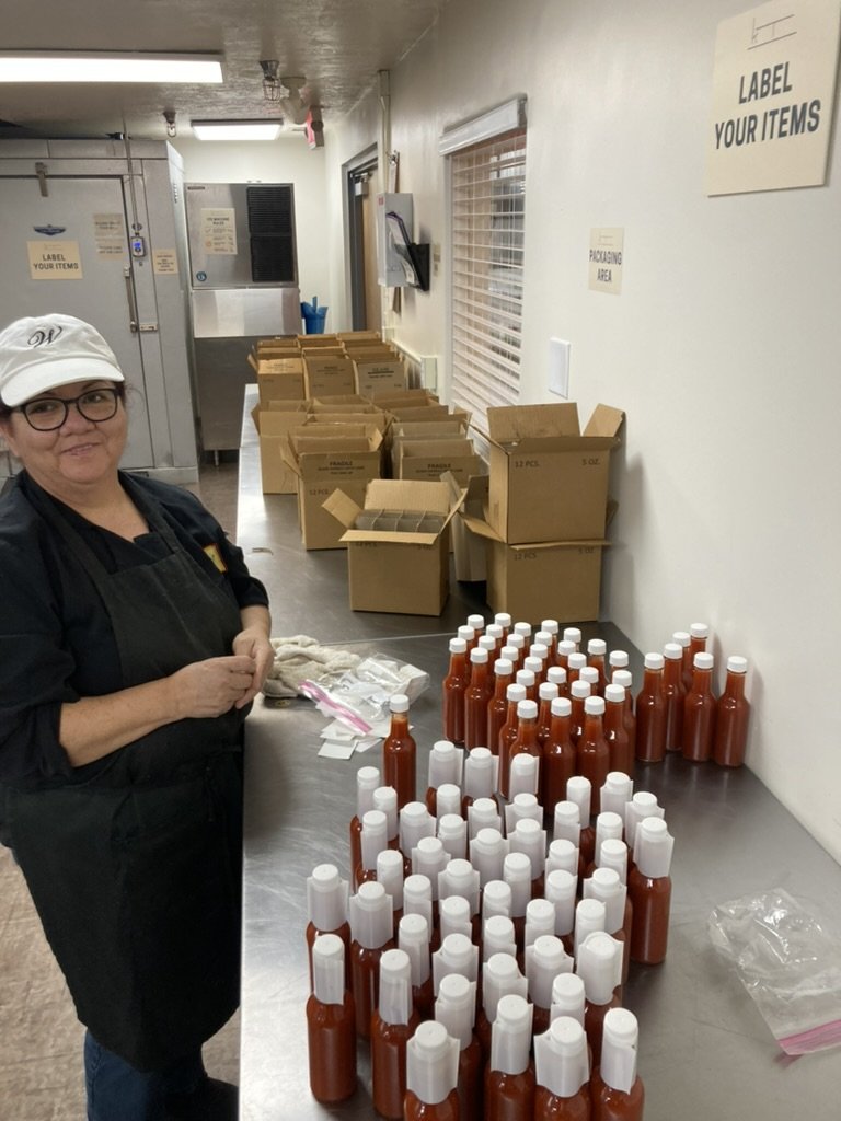 A woman in a white cap, glasses, black apron, and black shirt is smiling while standing next to a table with several bottles of hot sauce, with boxes and packaging supplies in the background.