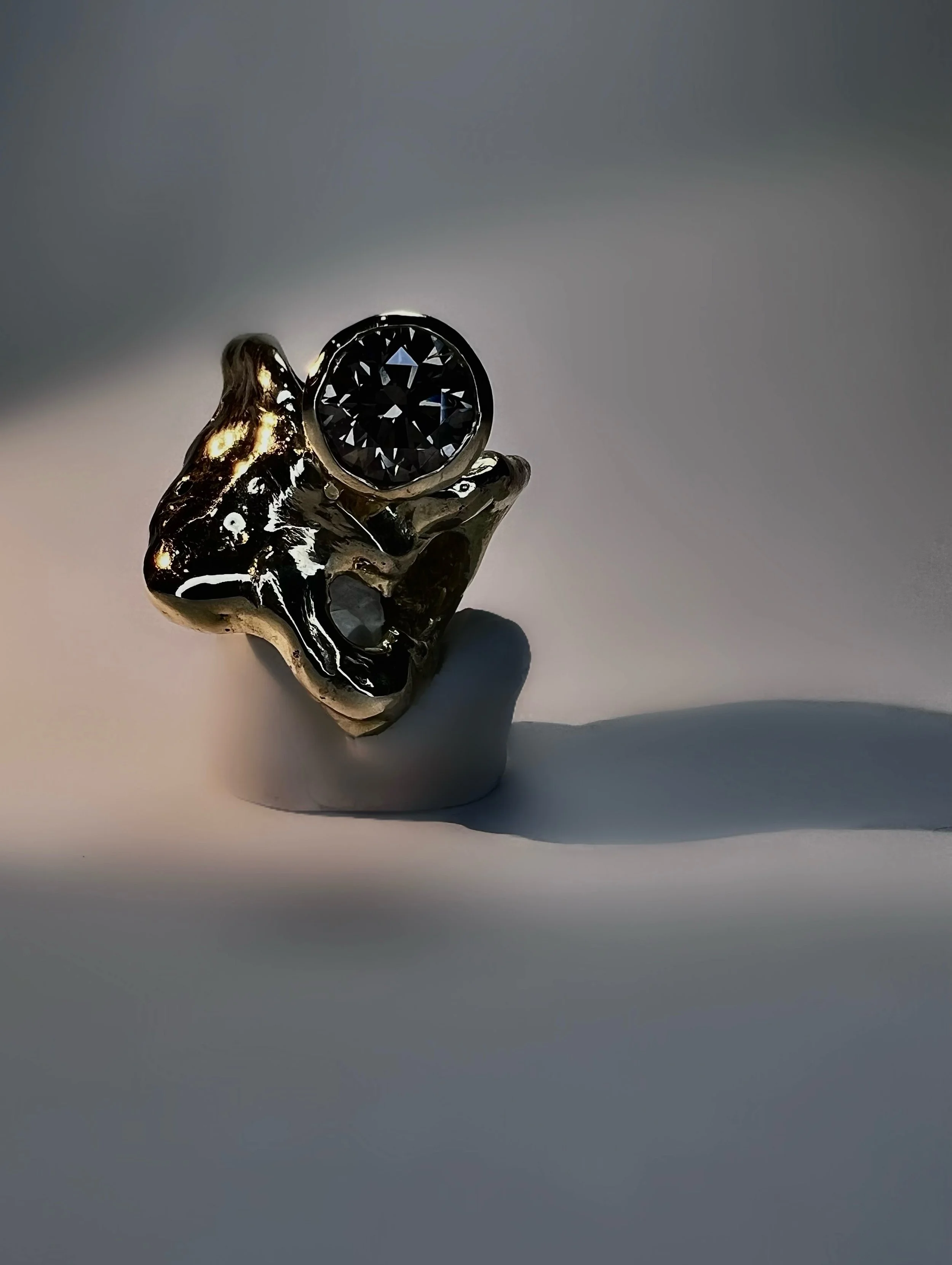 A silver ring with a large round black gemstone set in the center, held between fingers with the thumb and index finger visible. The background is plain and gray.