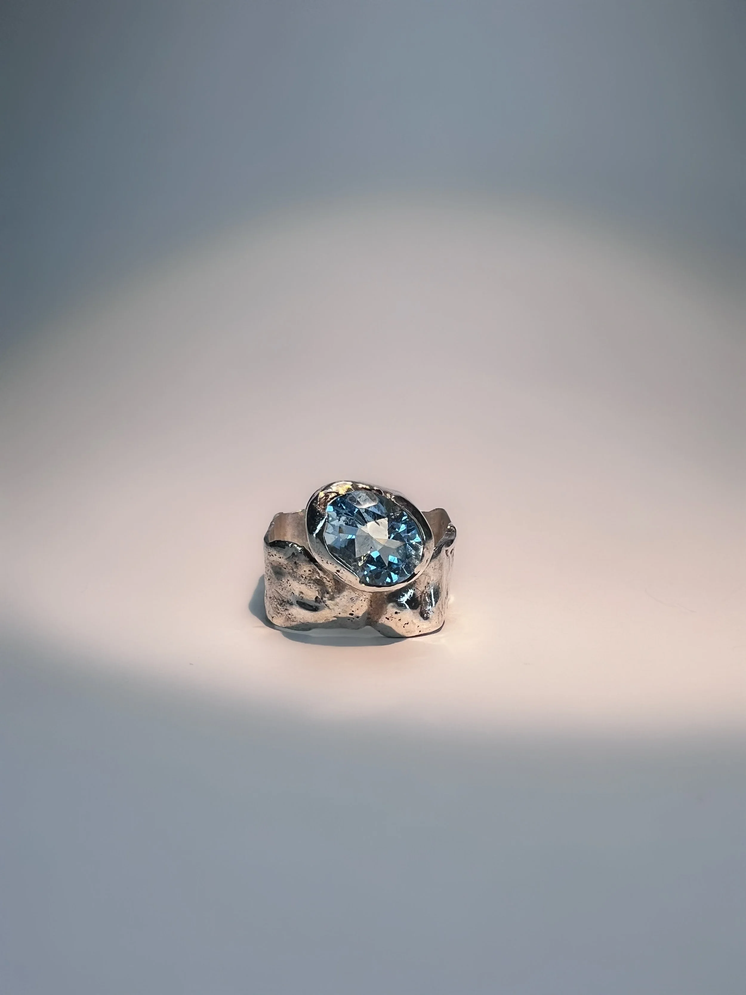 A silver ring with a large, oval-shaped blue gemstone set in the center, displayed on a white surface with a plain background.