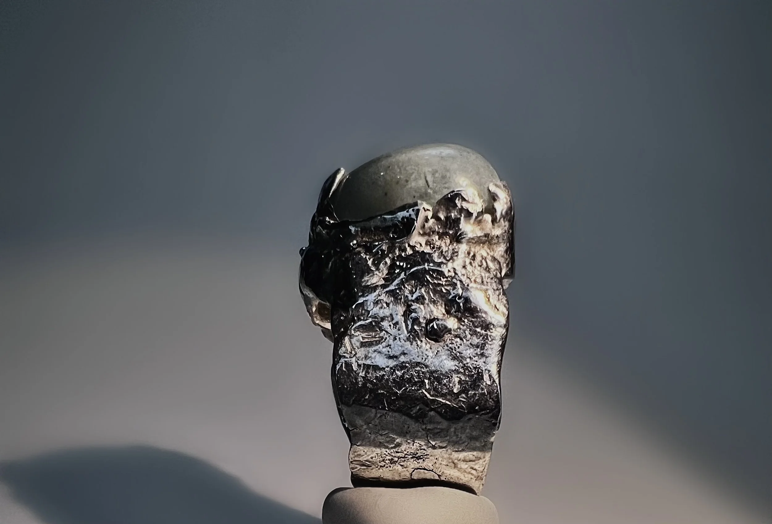 Close-up of a small, dark-colored, weathered rock balanced on a white pedestal against a plain, neutral background.