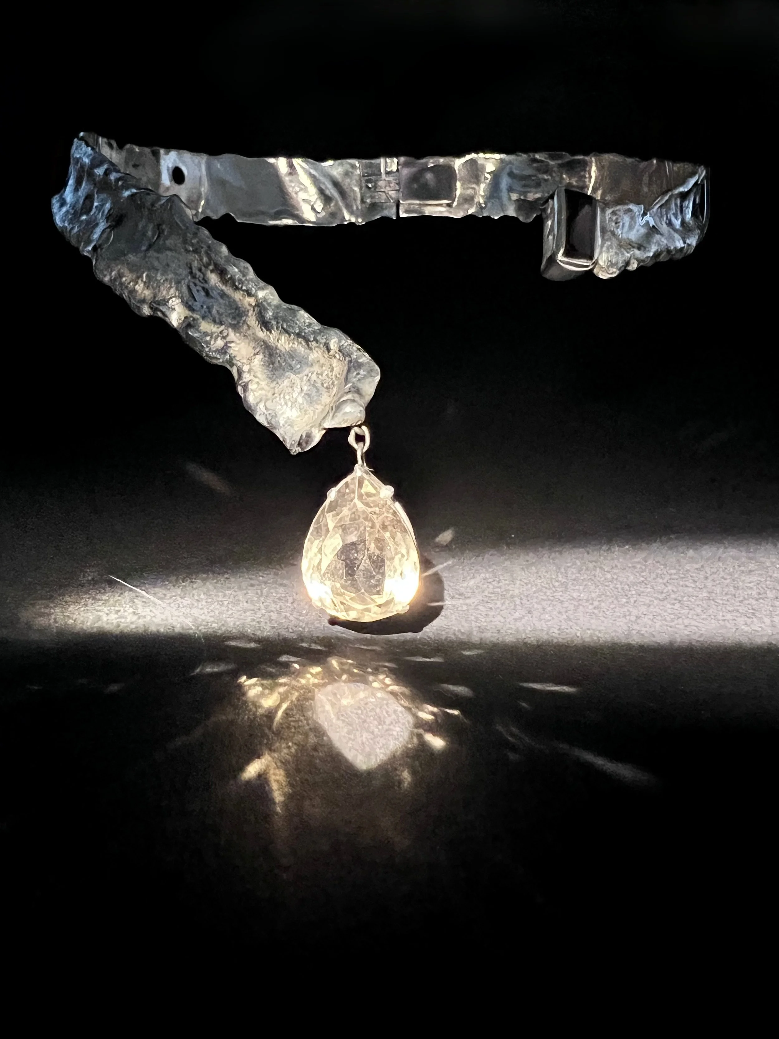 A metallic ring with textured, uneven surface and a water droplet hanging from it, illuminated against a black background.