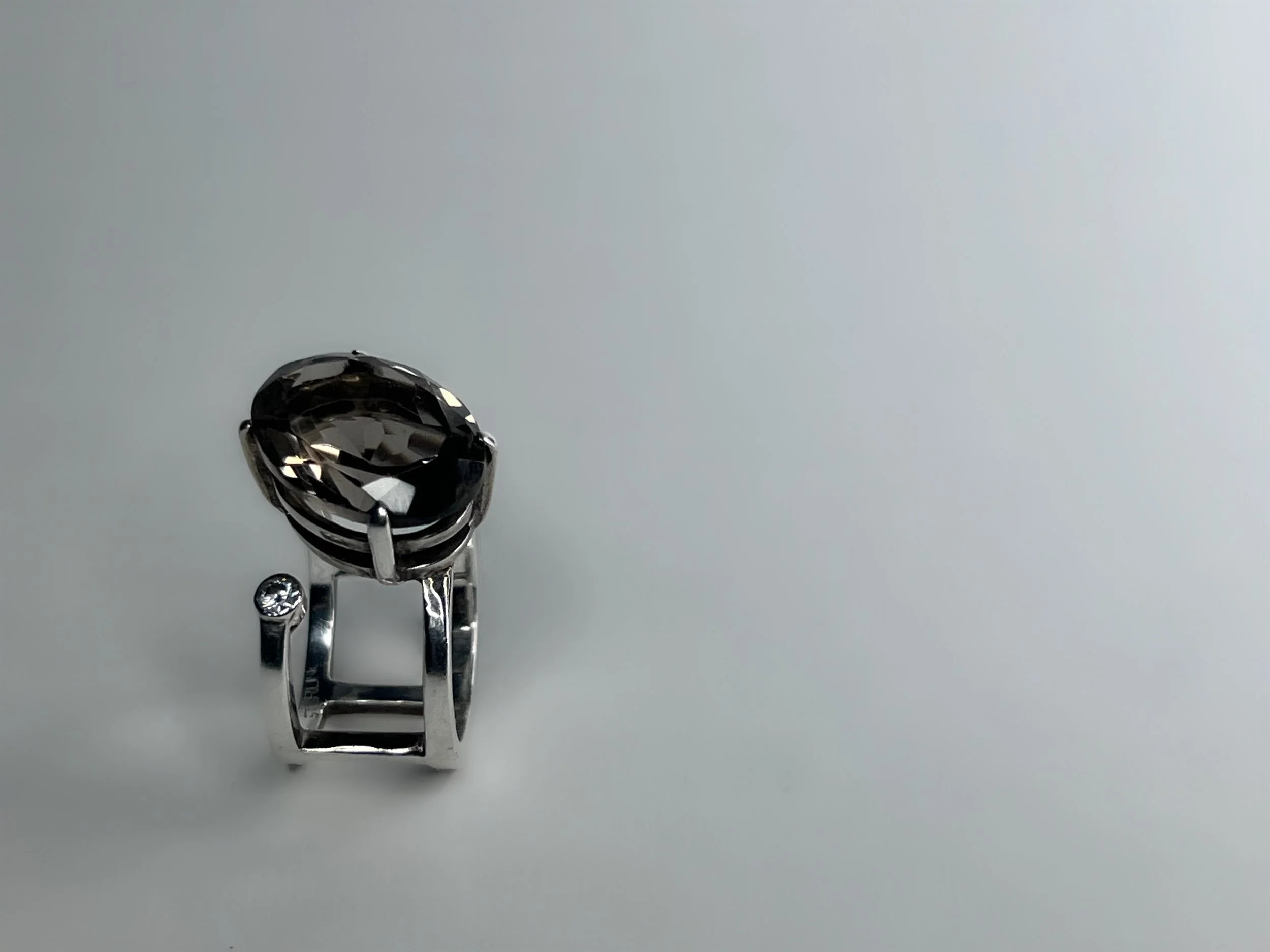 A silver ring with a large, dark, faceted gemstone mounted on a small metal stand, photographed against a plain white background.