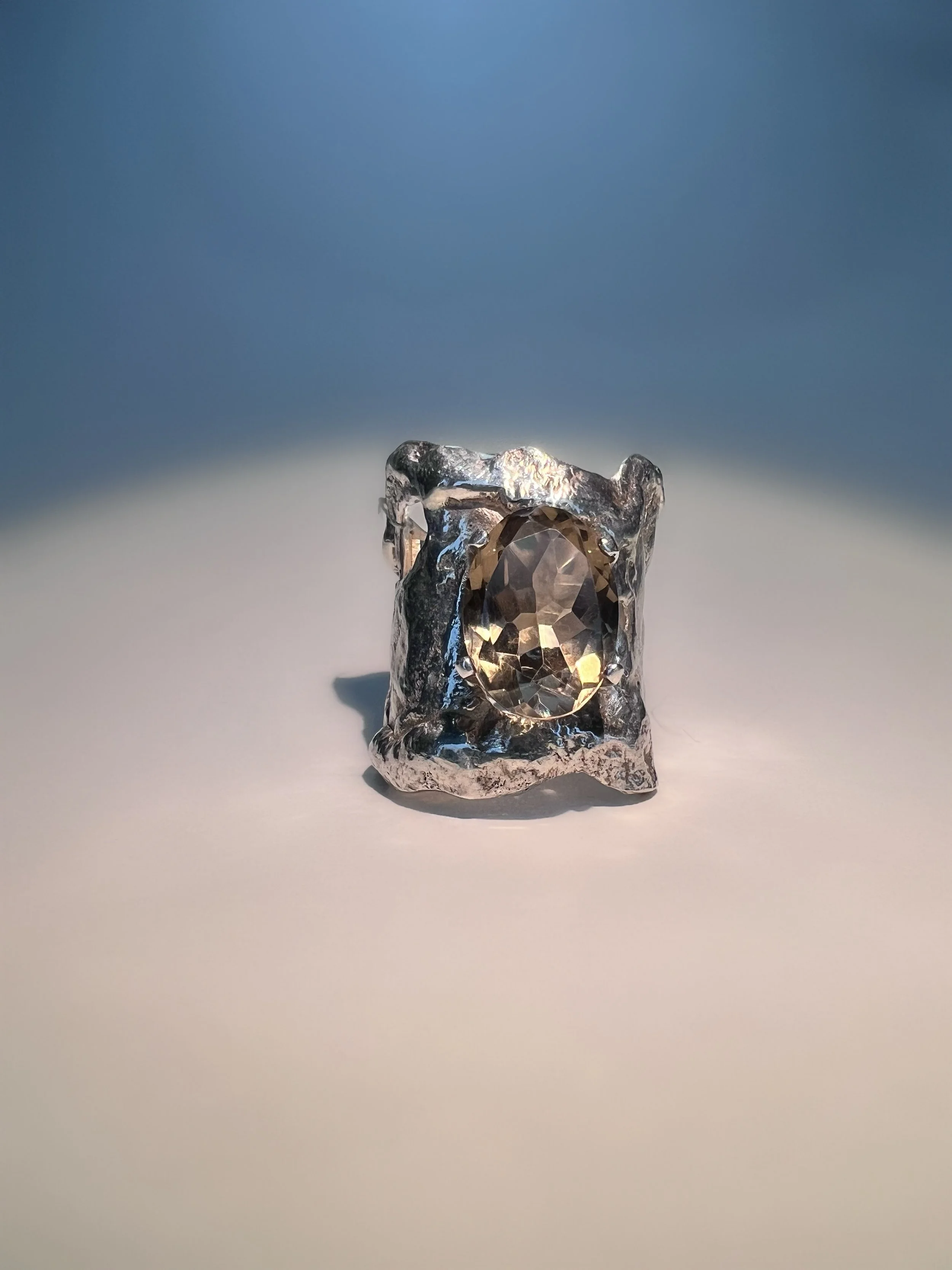 A silver ring with a large oval-shaped gemstone in the center, set on a silver textured band, against a gradient blue background.