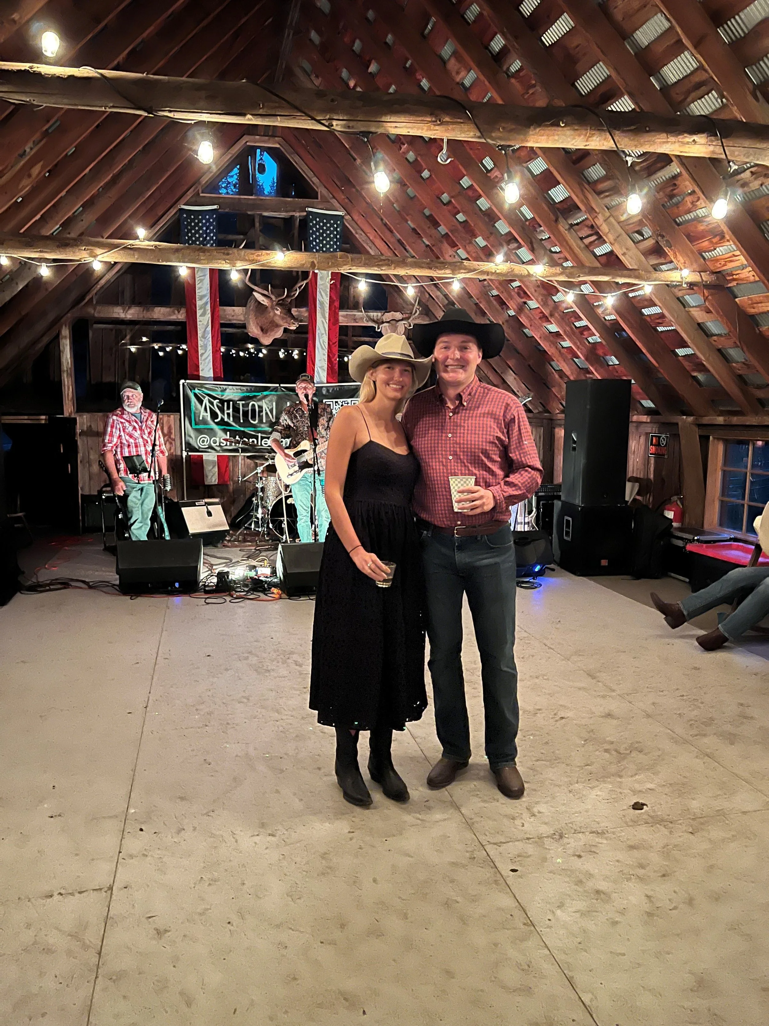 A man and woman dressed in country attire standing in front of a band on stage at a rustic indoor venue with wooden beams and American flag decorations.