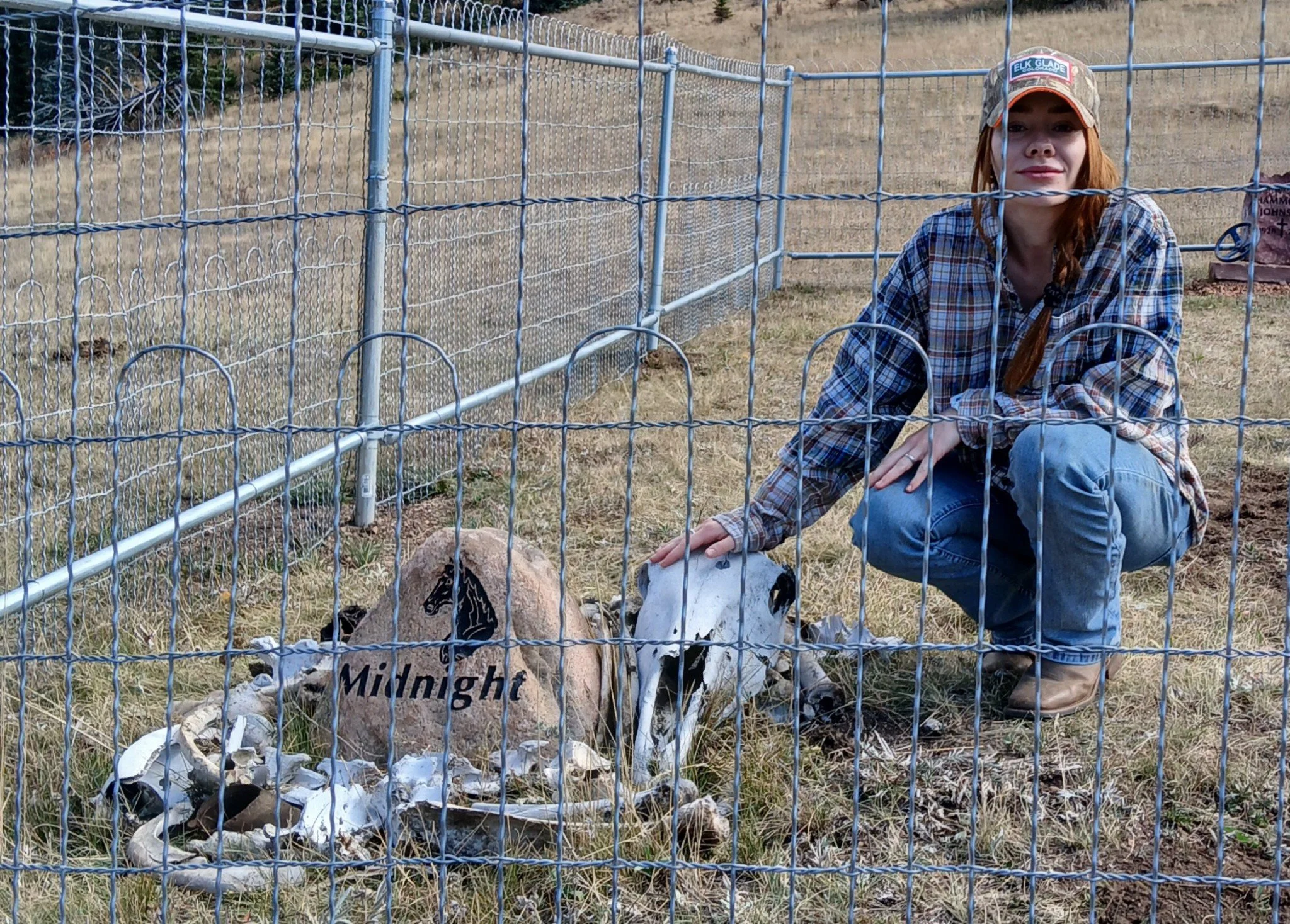 A woman crouching next to animal skulls and bones inside a fenced animal enclosure, with a wooden sign that has a horse illustration and the word 'Midnight' written on it.