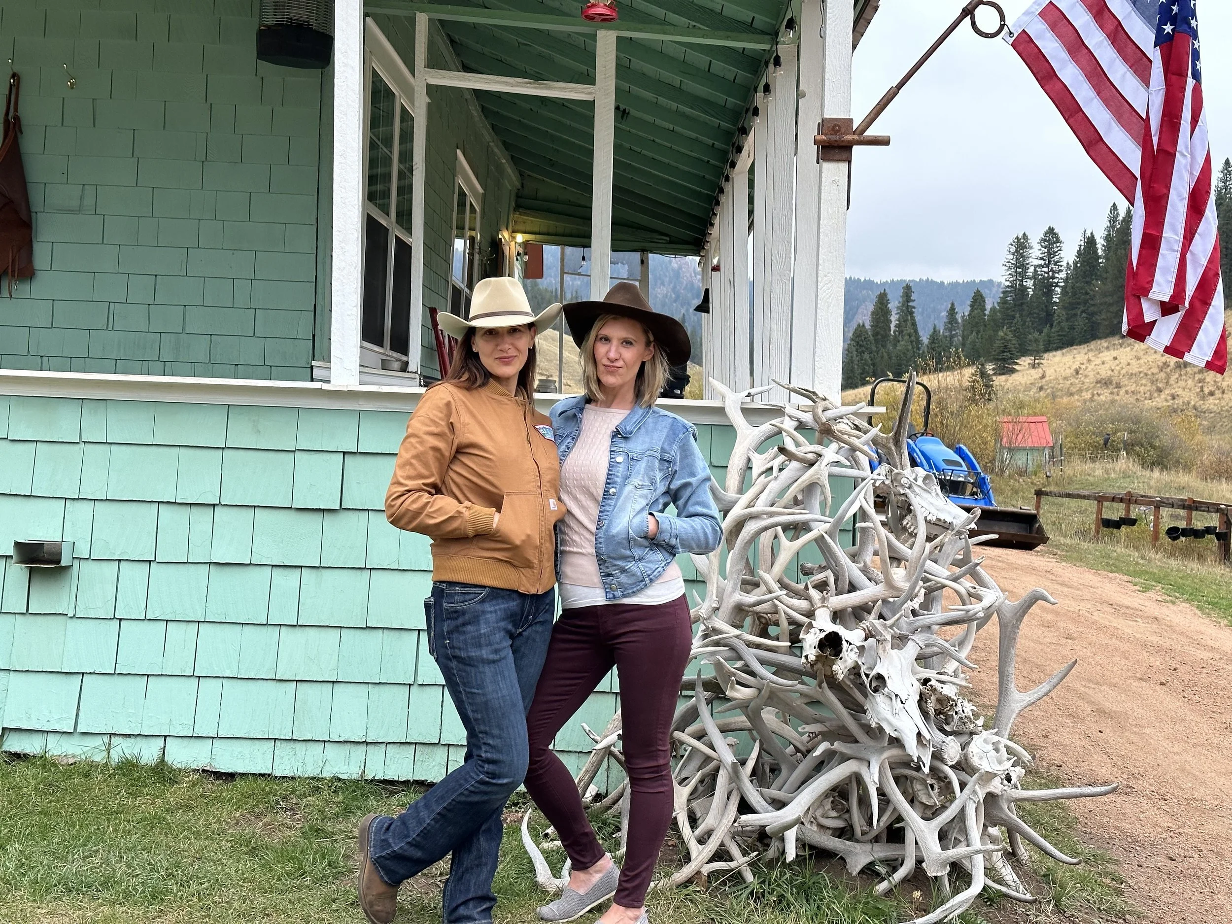 Two women wearing cowboy hats stand outdoors beside a pile of antlers and animal skulls, in front of a house with green shingle siding, a porch, and an American flag. The setting appears rural with hills and trees in the background.