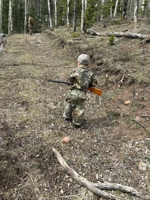 Child dressed in camouflage clothes holding a toy rifle, walking up a dirt trail in a wooded area.