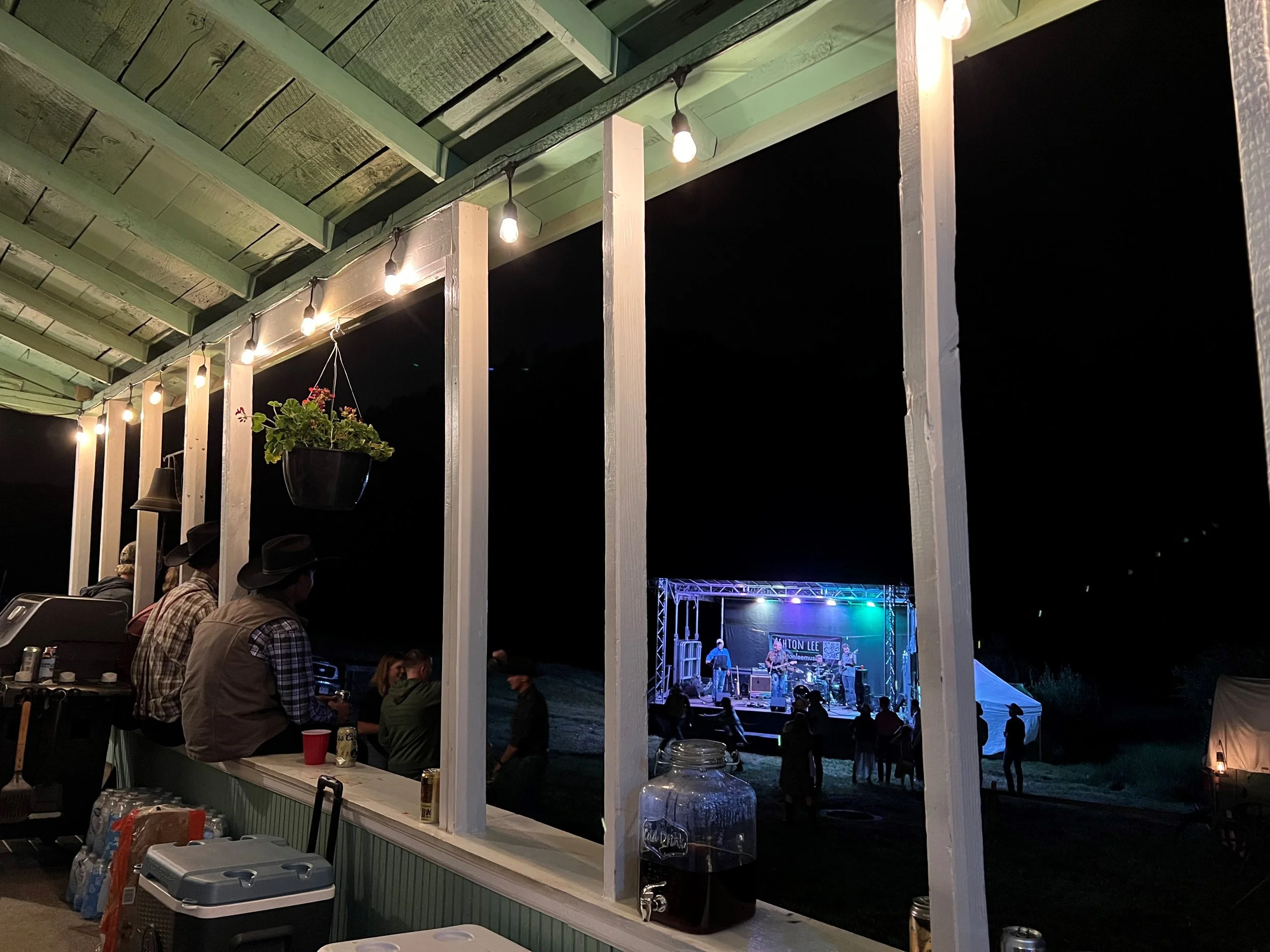 Nighttime outdoor concert scene viewed from a porch with string lights, a hanging plant, and a few people seated. In the distance, a live band plays on a stage with a sign reading 'Highton Lee,' and small groups of people are gathered nearby.