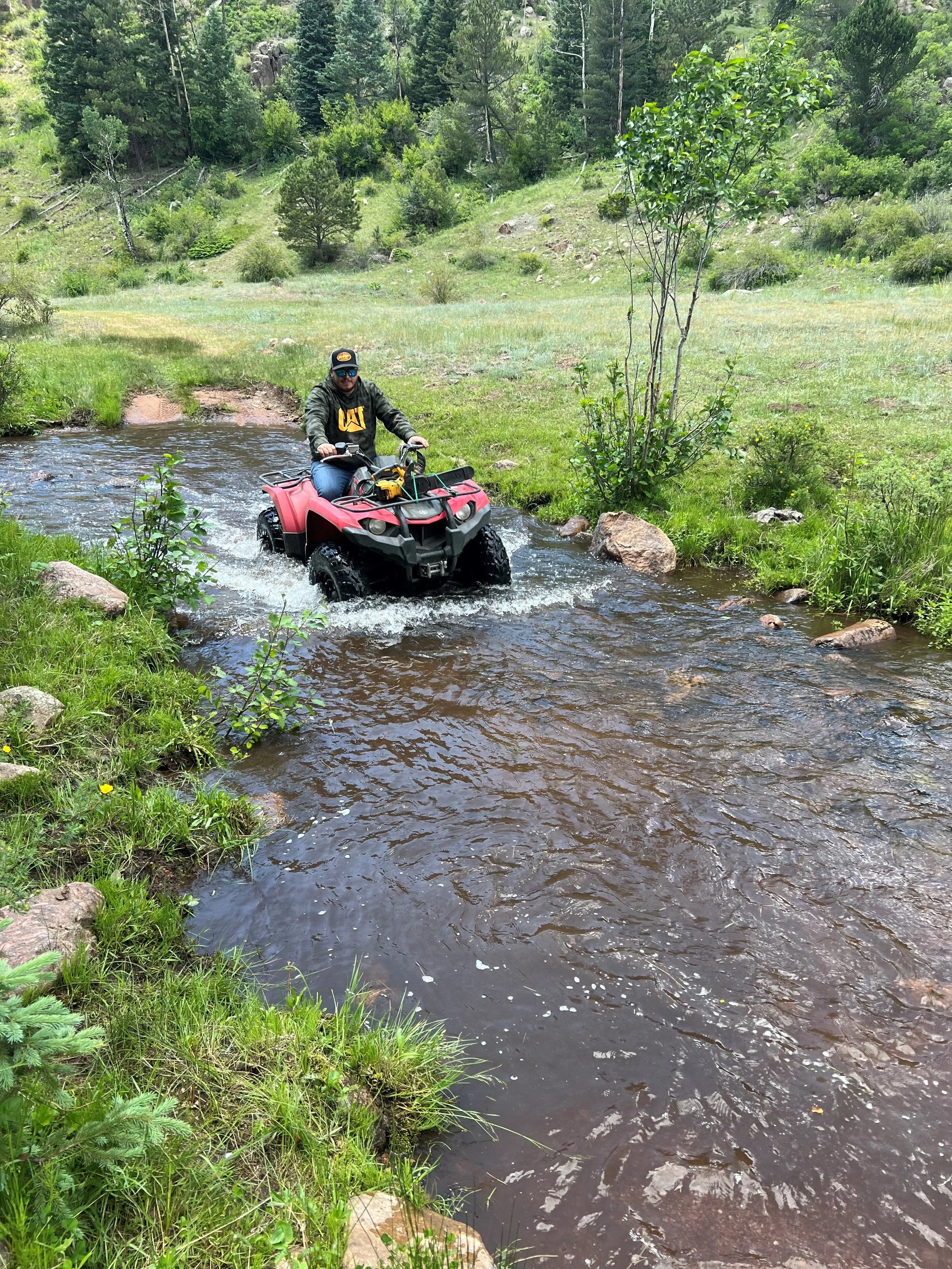 Man riding a red ATV across a shallow creek in a green forested landscape.