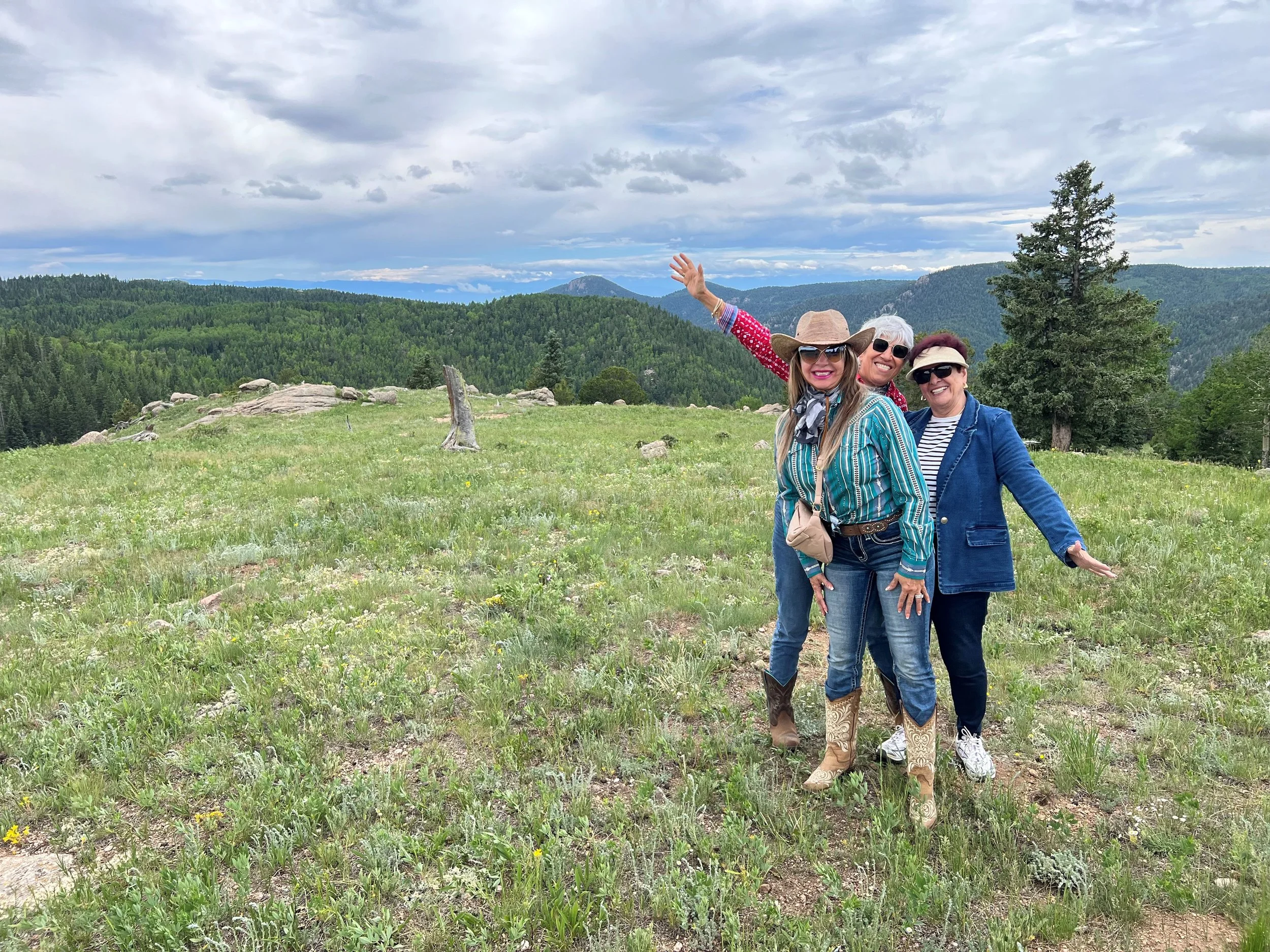 Three women smiling and posing in a grassy field with mountains and trees in the background, wearing casual outdoor clothing and sunglasses.