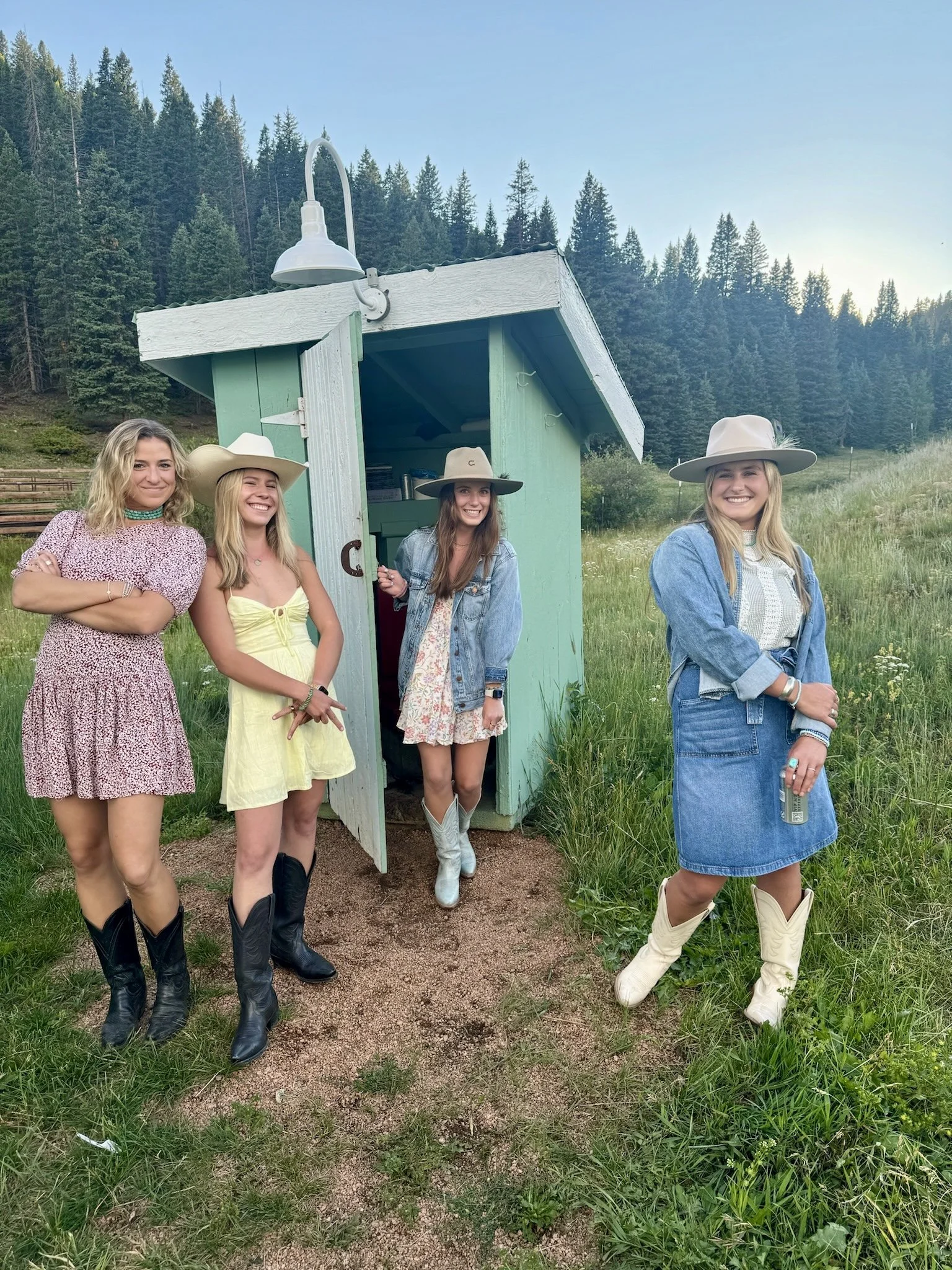 Four women standing outside in front of a small green building in a grassy outdoor setting with trees and mountains in the background. They are wearing cowboy hats and cowboy boots, smiling and posing for the photo.