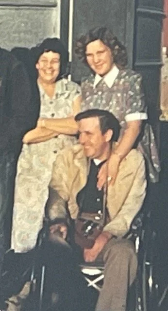 Two women standing behind a seated man with a camera around his neck, all smiling.