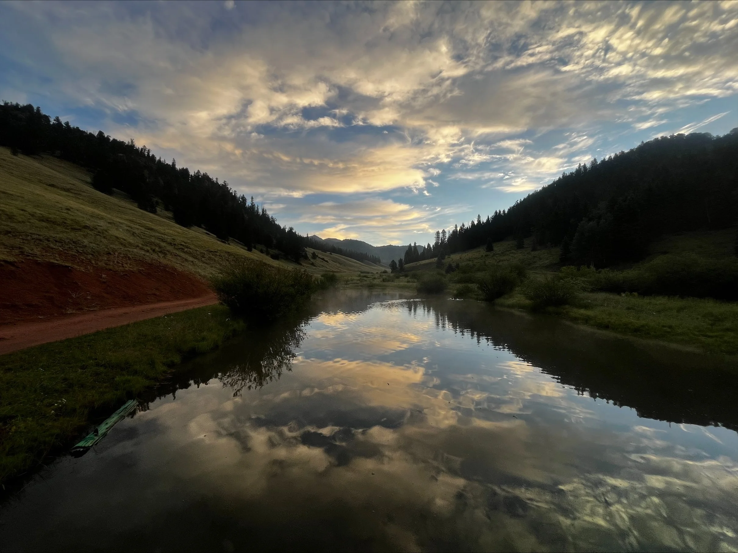 A peaceful river runs through a valley surrounded by hills and trees, with a partly cloudy sky reflecting on the water during sunset.