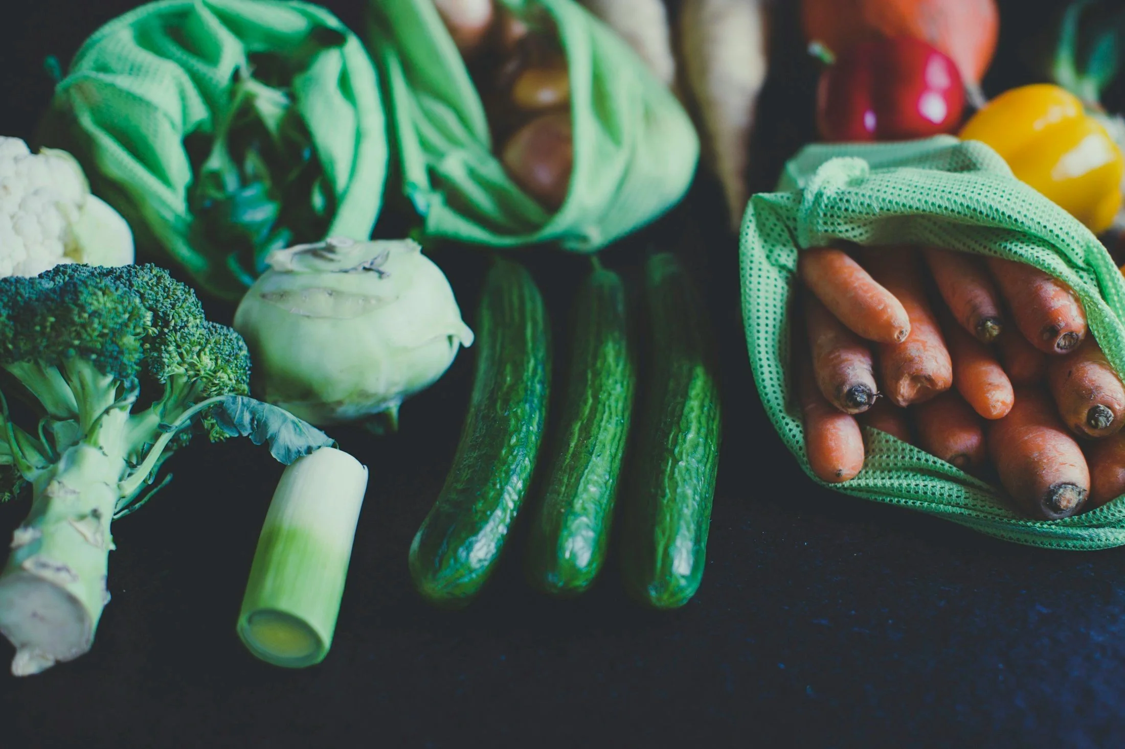 Fresh vegetables including broccoli, leeks, cucumbers, cauliflower, radicchio, carrots, and bell peppers displayed on a dark surface.