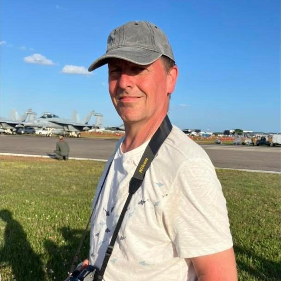 A man in a gray cap and light-colored t-shirt stands on grass with military aircraft in the background at an airshow, holding a Nikon camera strap.