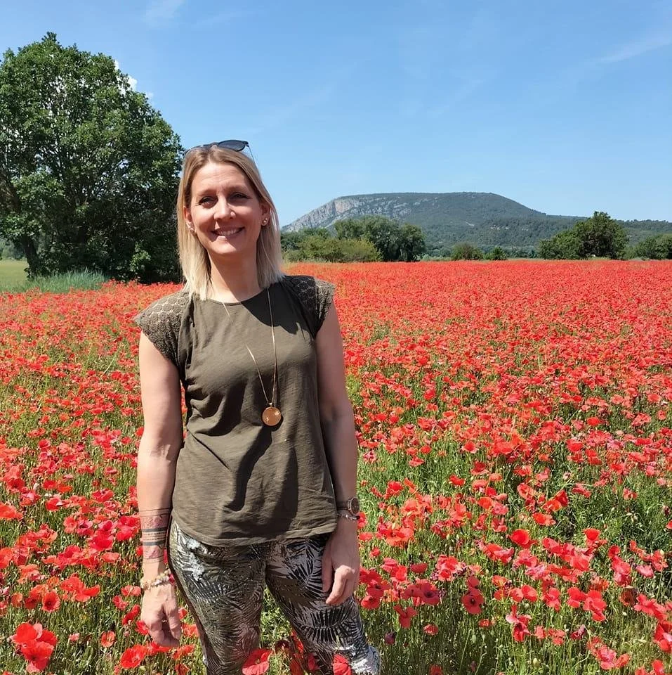 Une femme souriante debout dans un champ de coquelicots rouges avec un arbre, une montagne et un ciel bleu en arrière-plan.