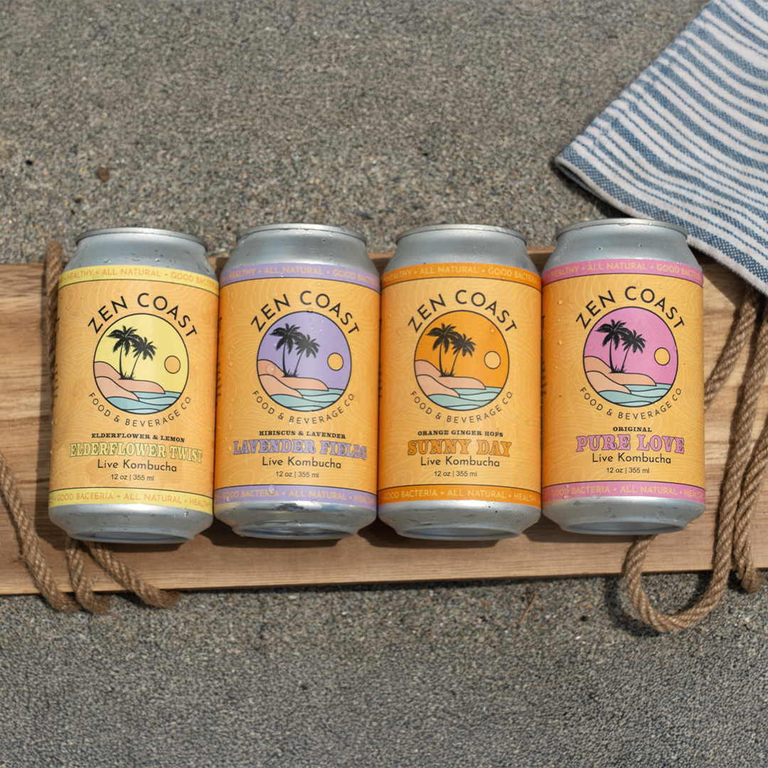 Four cans of Zen Coast live kombucha on a wooden board on sandy beach, each with different flavors: Elderflower Lemon, Lavender Fields, Sunny Day, Pure Love, with a striped towel partially visible to the right.