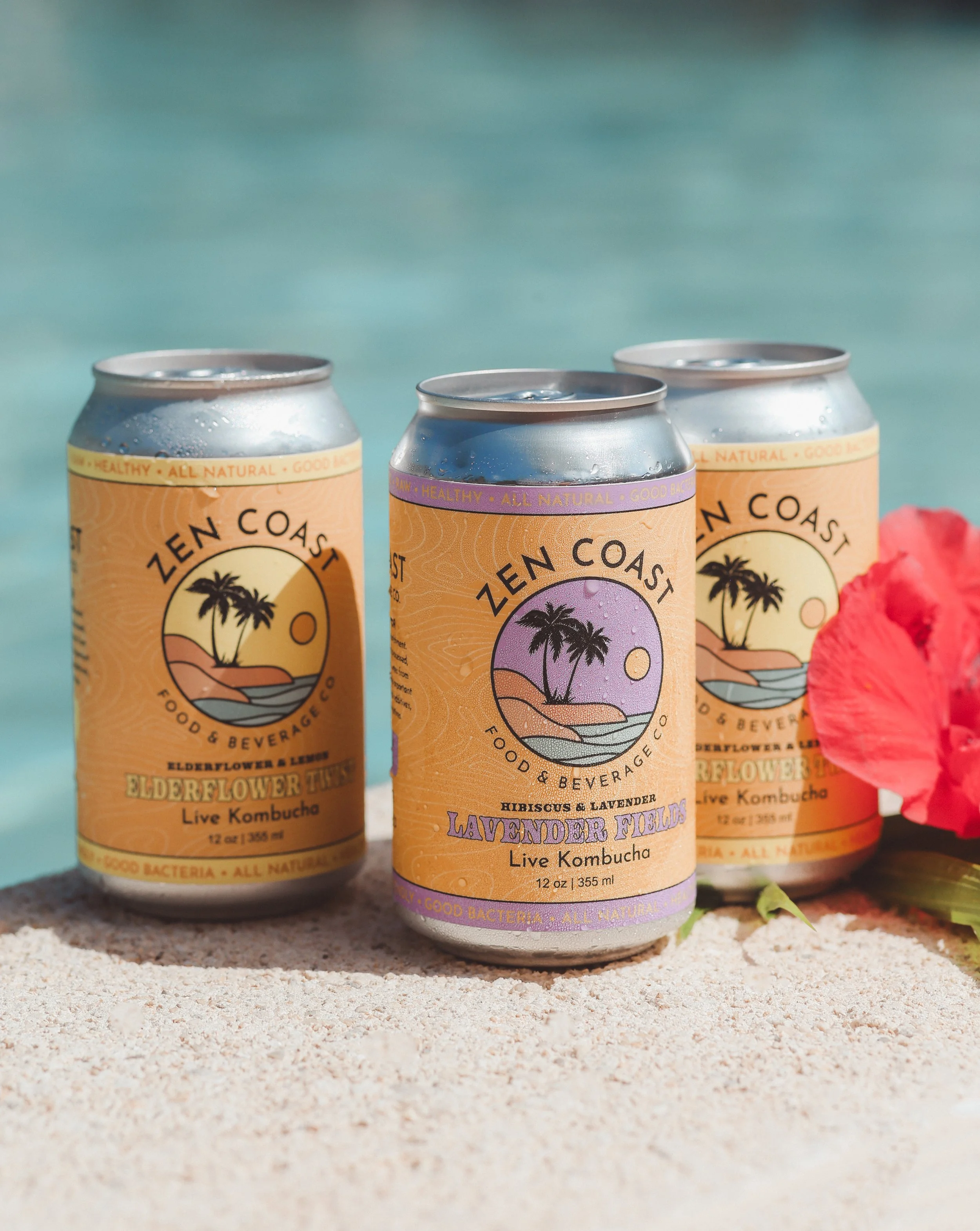 Three cans of Zen Coast live kombucha flavored Elderflower Lemon, Hibiscus & Lavender, and Lavender Fields, placed on sandy ground with a turquoise background and pink flower petal.